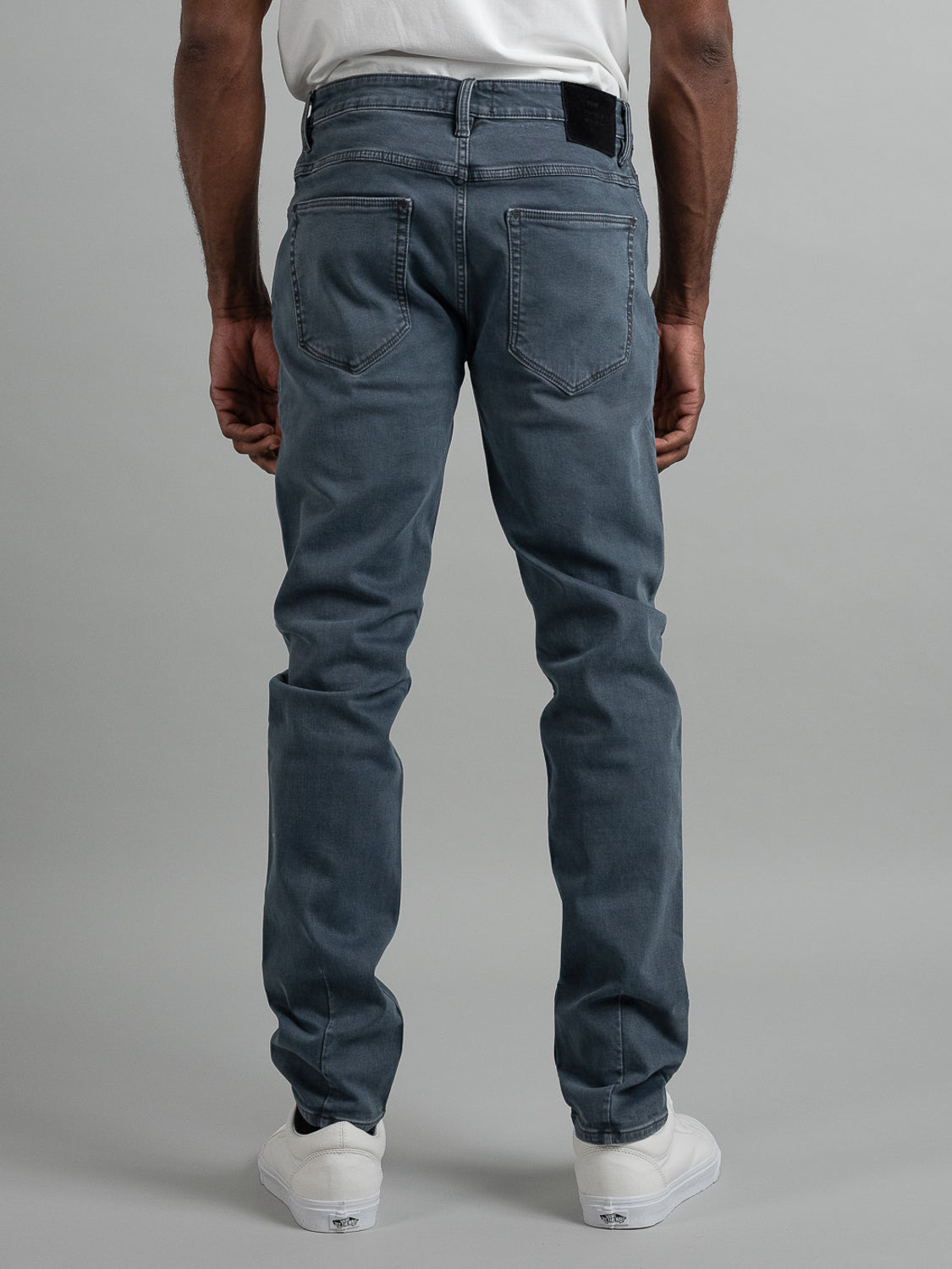 The lower half of a person is shown facing away, wearing Neuw's Lou Slim Mercury faded blue stretch denim jeans, a white t-shirt, and white sneakers against a plain gray background.