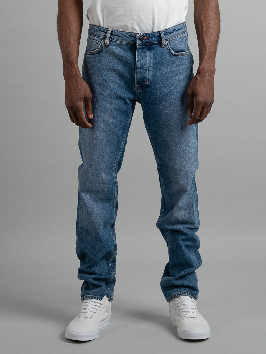 A person wearing Neuw Ray Straight Shock blue jeans, a white t-shirt, and white sneakers stands against a plain gray background, with only the lower torso and legs visible.