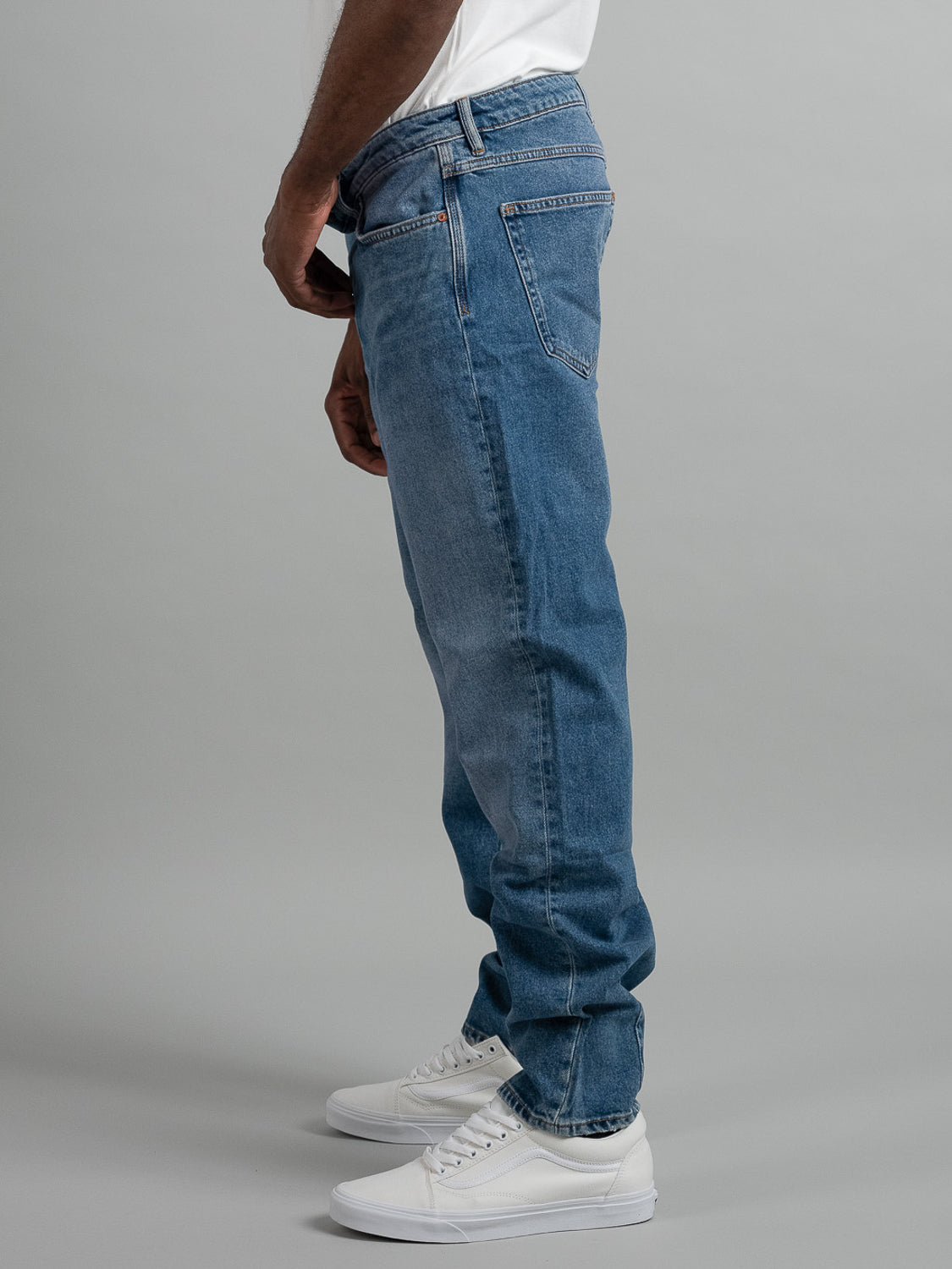A person stands sideways wearing Neuw Ray Straight Shock blue jeans, a white t-shirt, and white sneakers against a plain gray background.