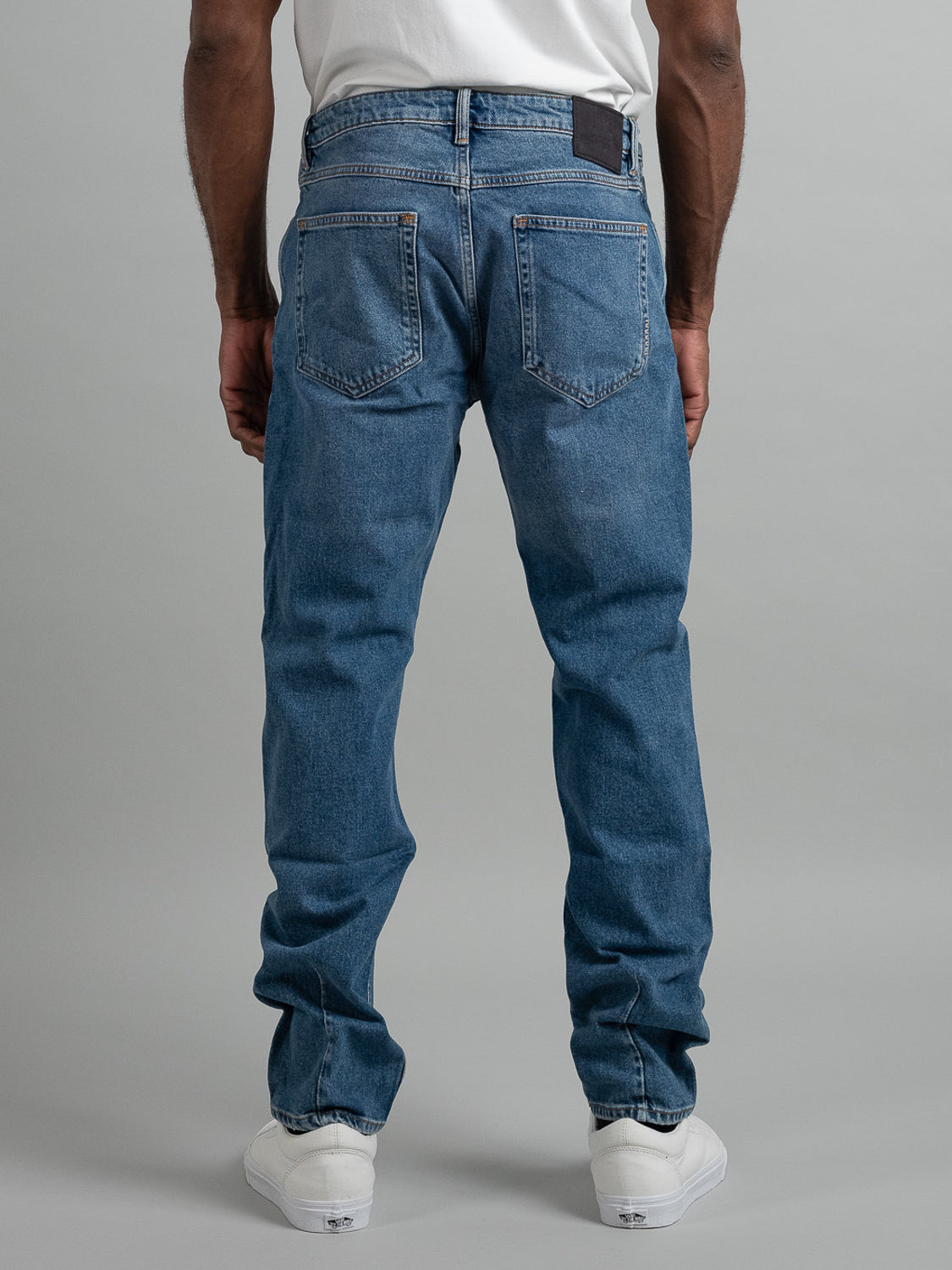 A person in a white shirt, white sneakers, and Neuw Ray Straight Shock jeans stands facing away from the camera, highlighting the back of the jeans against a plain gray background.