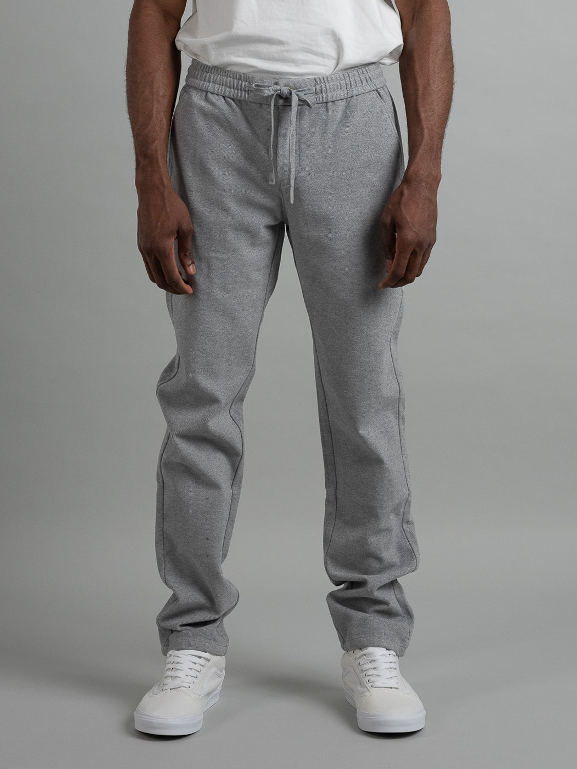 A person wears a white t-shirt and Brighton Brushed Polyester Rayon Pant from ID Menswear in light gray, featuring an elastic waist, against a gray backdrop. Theyre wearing white sneakers, highlighting the sleek design of the joggers lower half.
