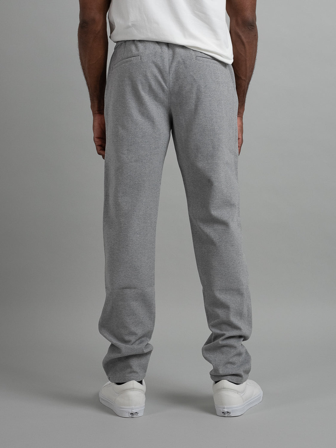 A person is standing against a plain gray background wearing Brighton Brushed Polyester Rayon Pants by ID Menswear in light gray and a white T-shirt, facing away to show the back. The look is completed with white sneakers.