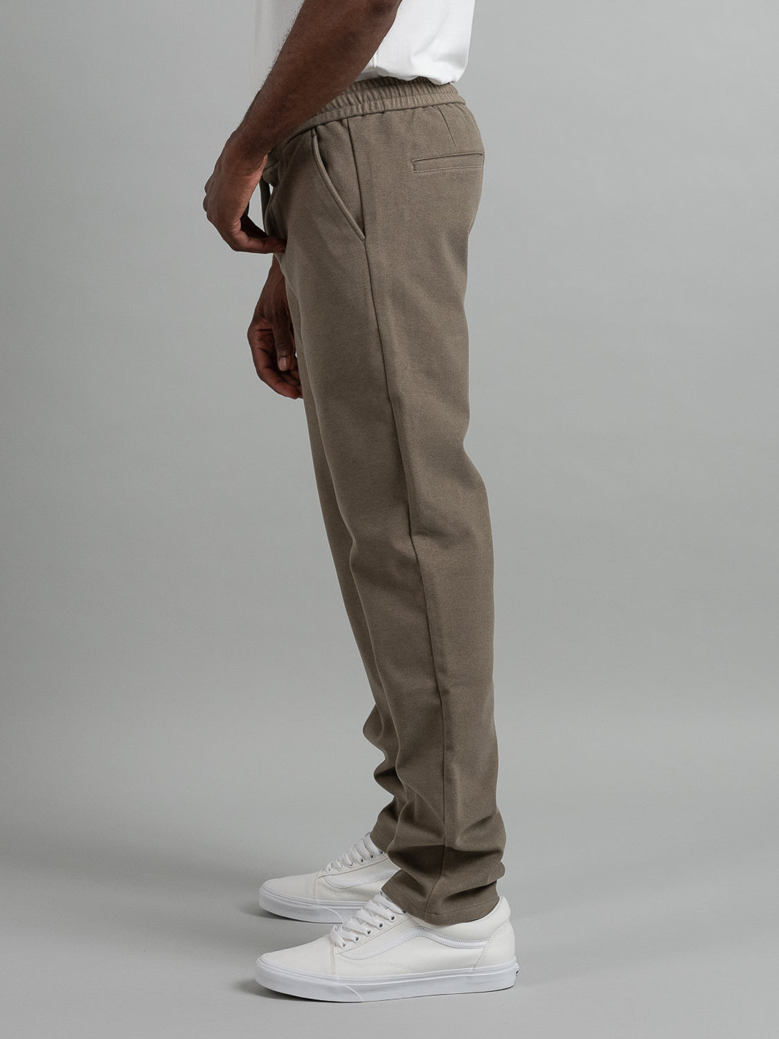 Side view of a person wearing Brighton Brushed Polyester Rayon Pants by ID Menswear in olive-green, paired with white sneakers against a light gray background. The white shirt is partially visible as their hands rest by their sides.