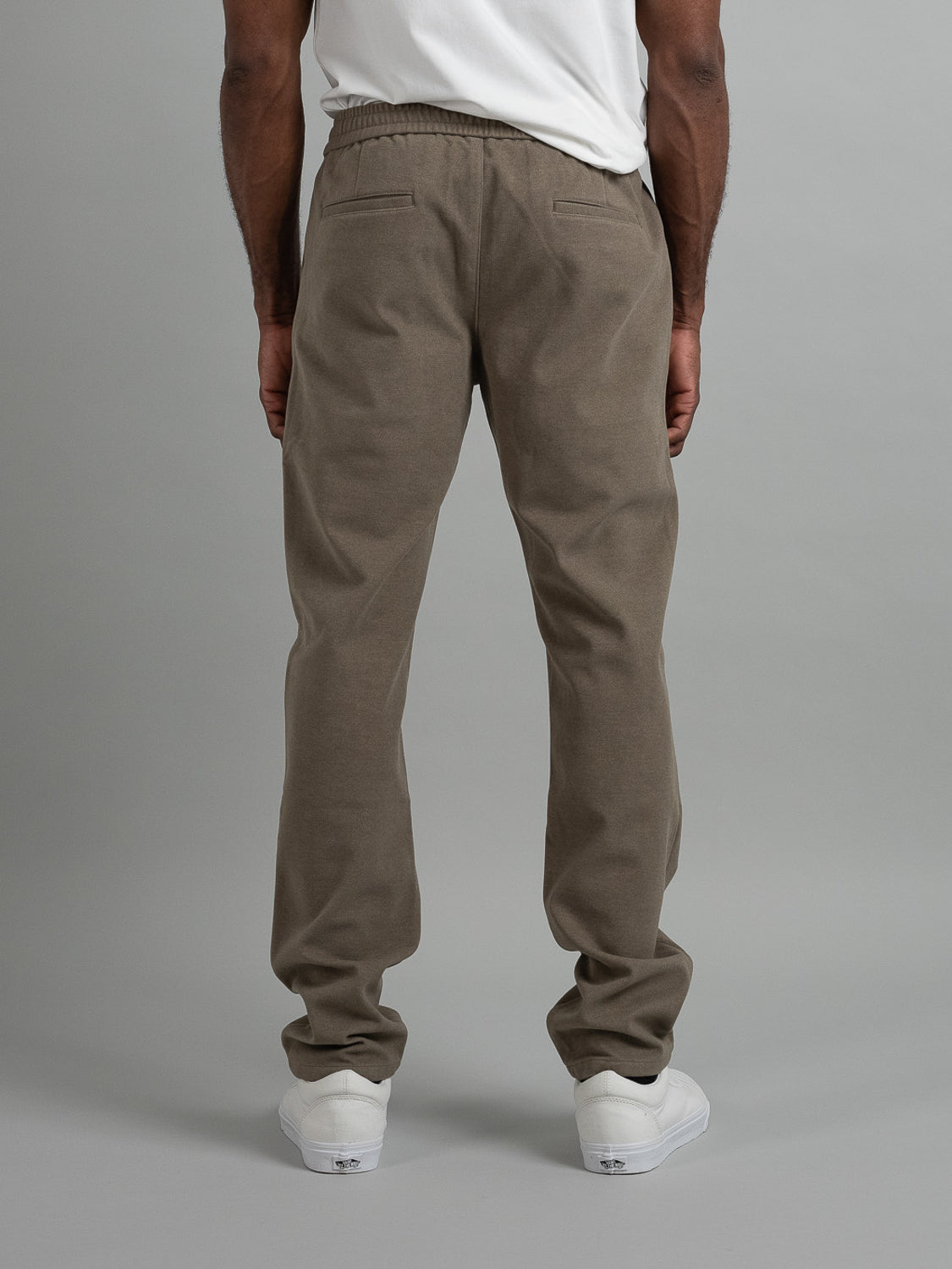 A person, seen from behind against a gray backdrop, is wearing an ID Menswear white shirt with Brighton Brushed Polyester Rayon Pants in olive green and white sneakers, their arms relaxed at their sides.