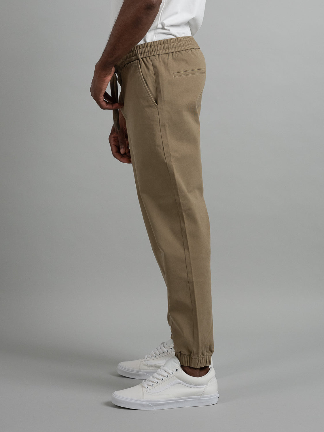 A side view showcases ID Menswears Dyker Cotton Twill Drawstring Jogger in beige, featuring an elastic waistband and ankle cuffs, paired with white sneakers and a crisp white shirt against a neutral background.