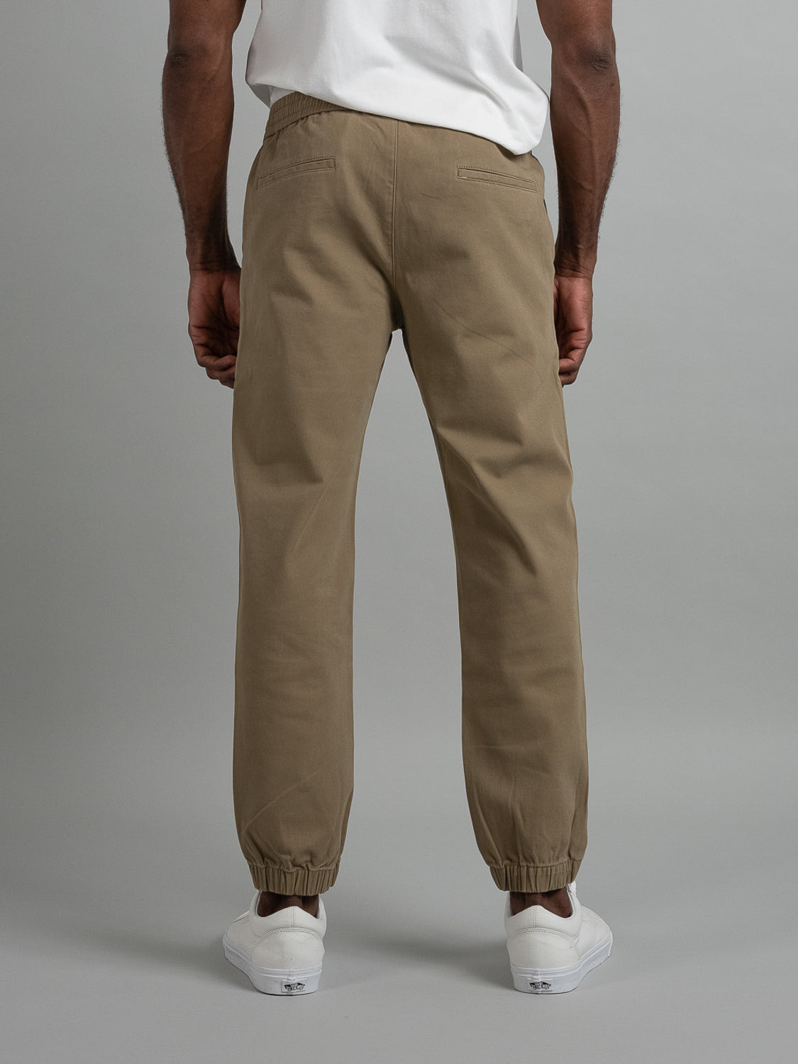 Rear view of a person in a white T-shirt, Dyker Cotton Twill Drawstring Jogger by ID Menswear with elastic cuffs, and white sneakers, standing on a gray background.