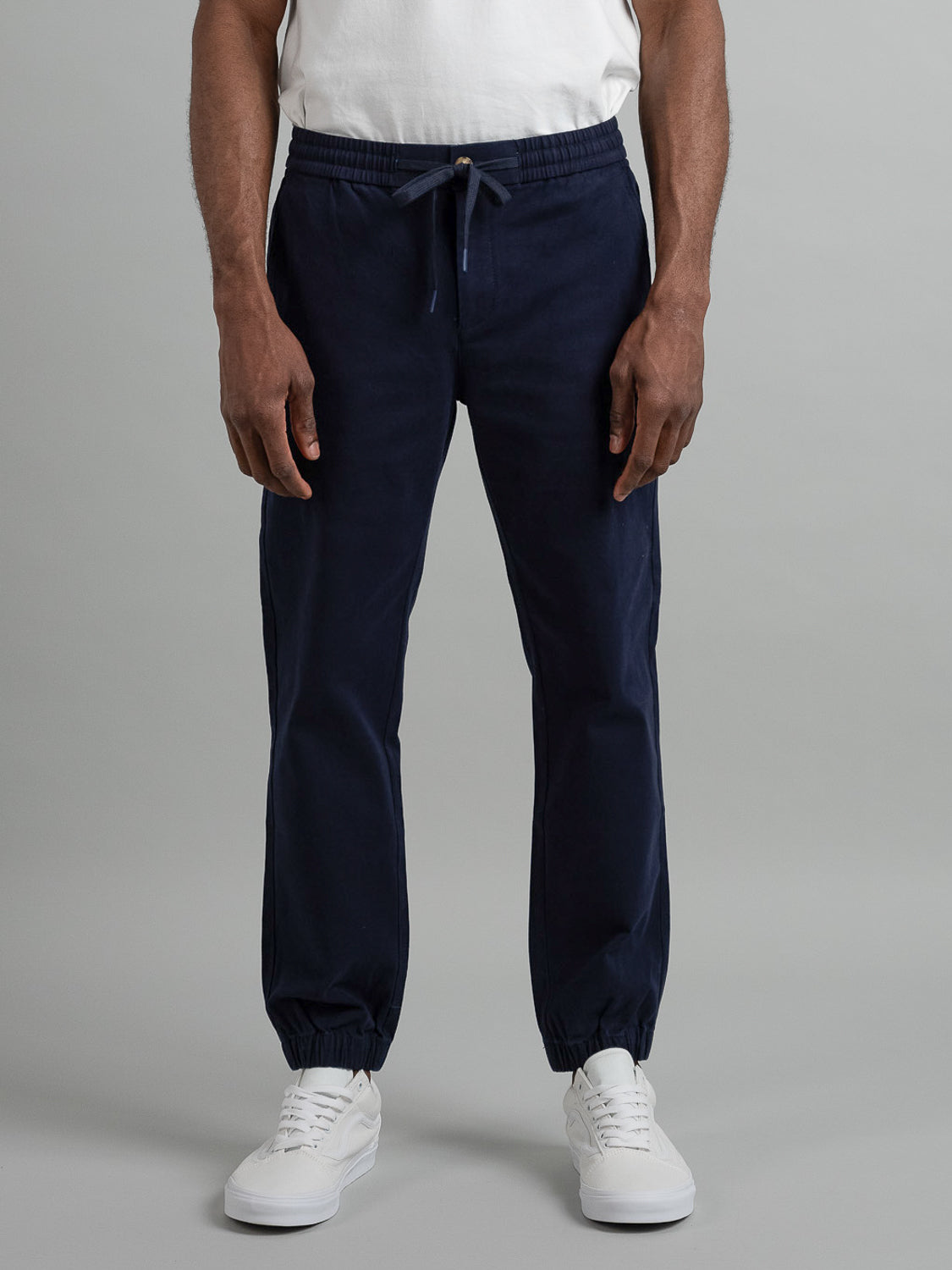 Someone is wearing the Dyker Cotton Twill Drawstring Jogger by ID Menswear, featuring elastic cuffs and a drawstring waist, paired with a white T-shirt and sneakers against a plain gray background.