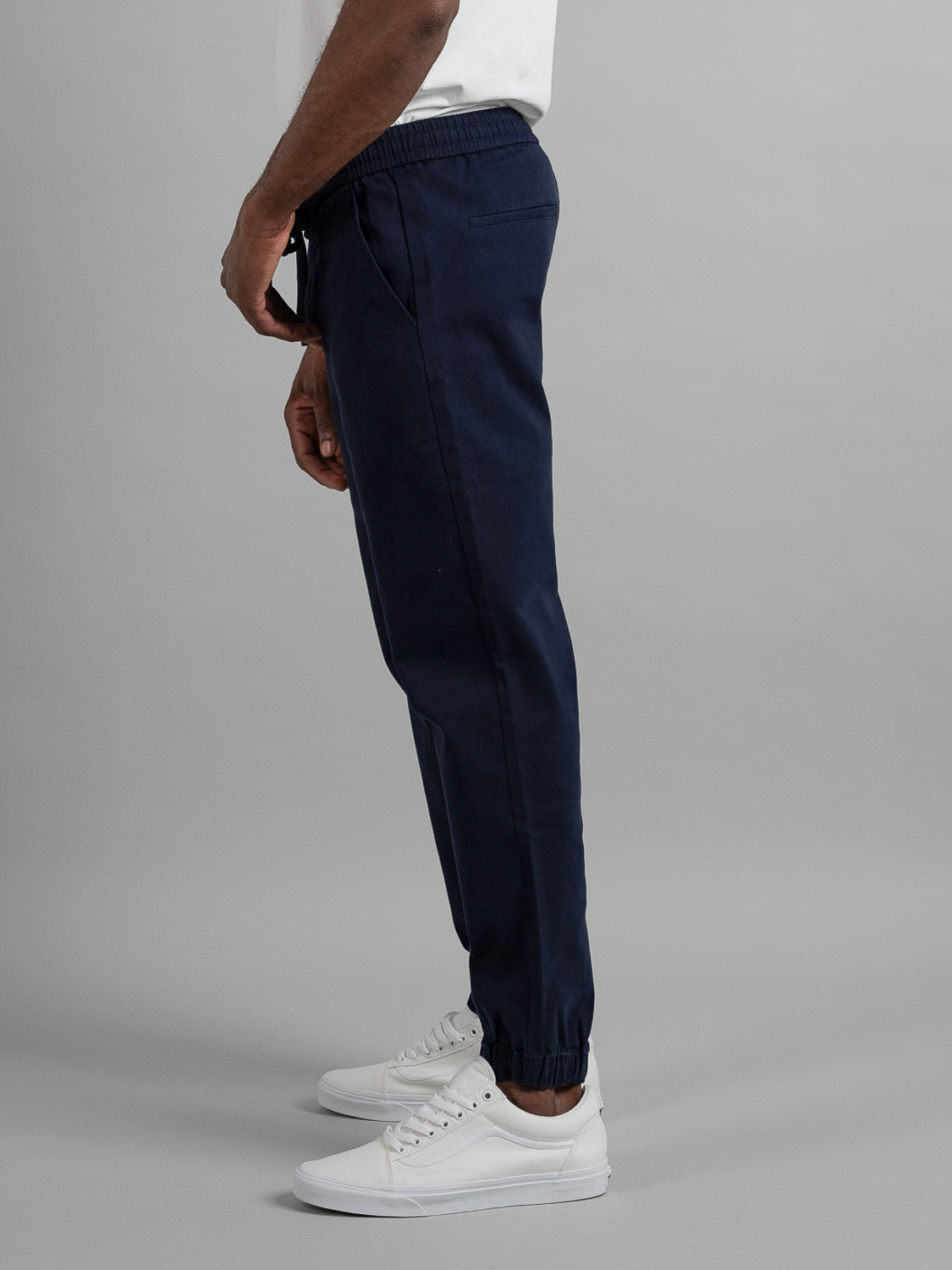 Side view of a person in ID Menswears Dyker Cotton Twill Drawstring Jogger with white sneakers against a gray background, highlighting the clothing and footwear.