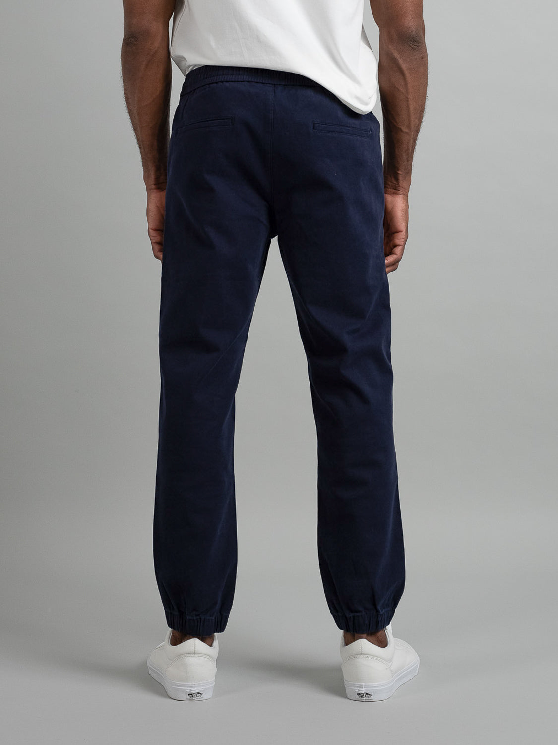 An individual stands back to the camera donning ID Menswears Dyker Cotton Twill Drawstring Jogger in dark blue, paired with white shoes. The joggers elastic waist adds comfort against a plain light gray backdrop.