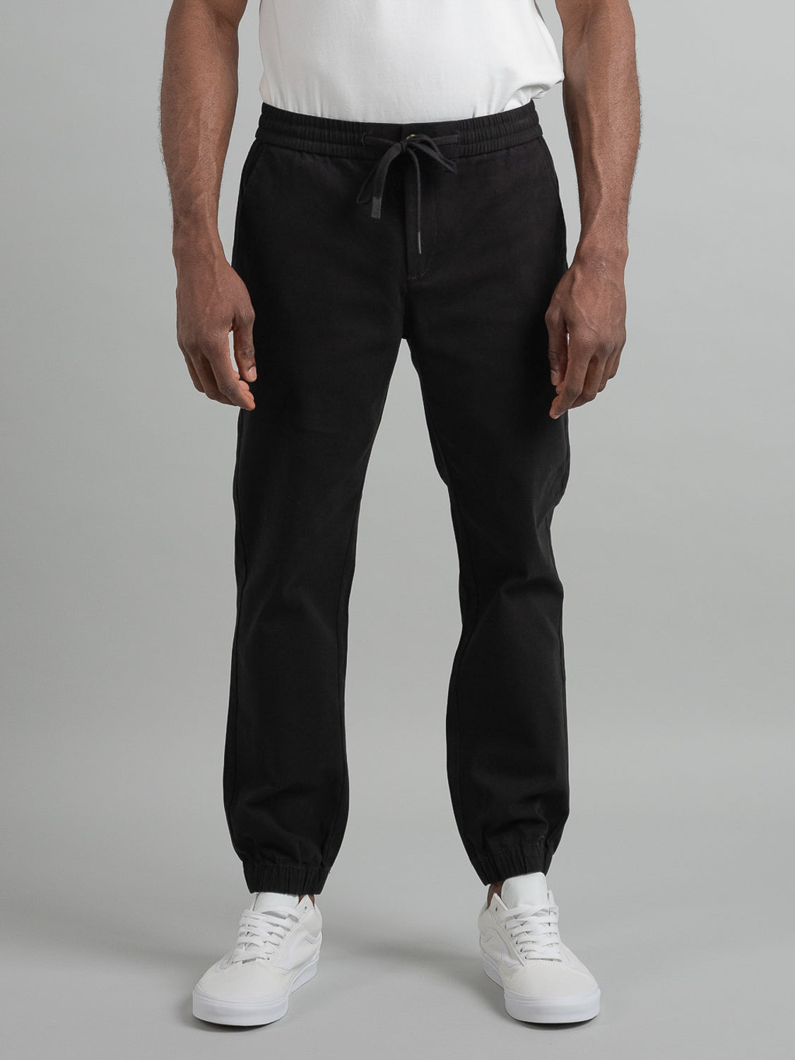 A person dressed in a white t-shirt, ID Menswears Dyker Cotton Twill Drawstring Jogger with an elastic waist in black, and white sneakers stands against a gray backdrop. Their arms are relaxed at their sides, exuding an effortlessly casual look.
