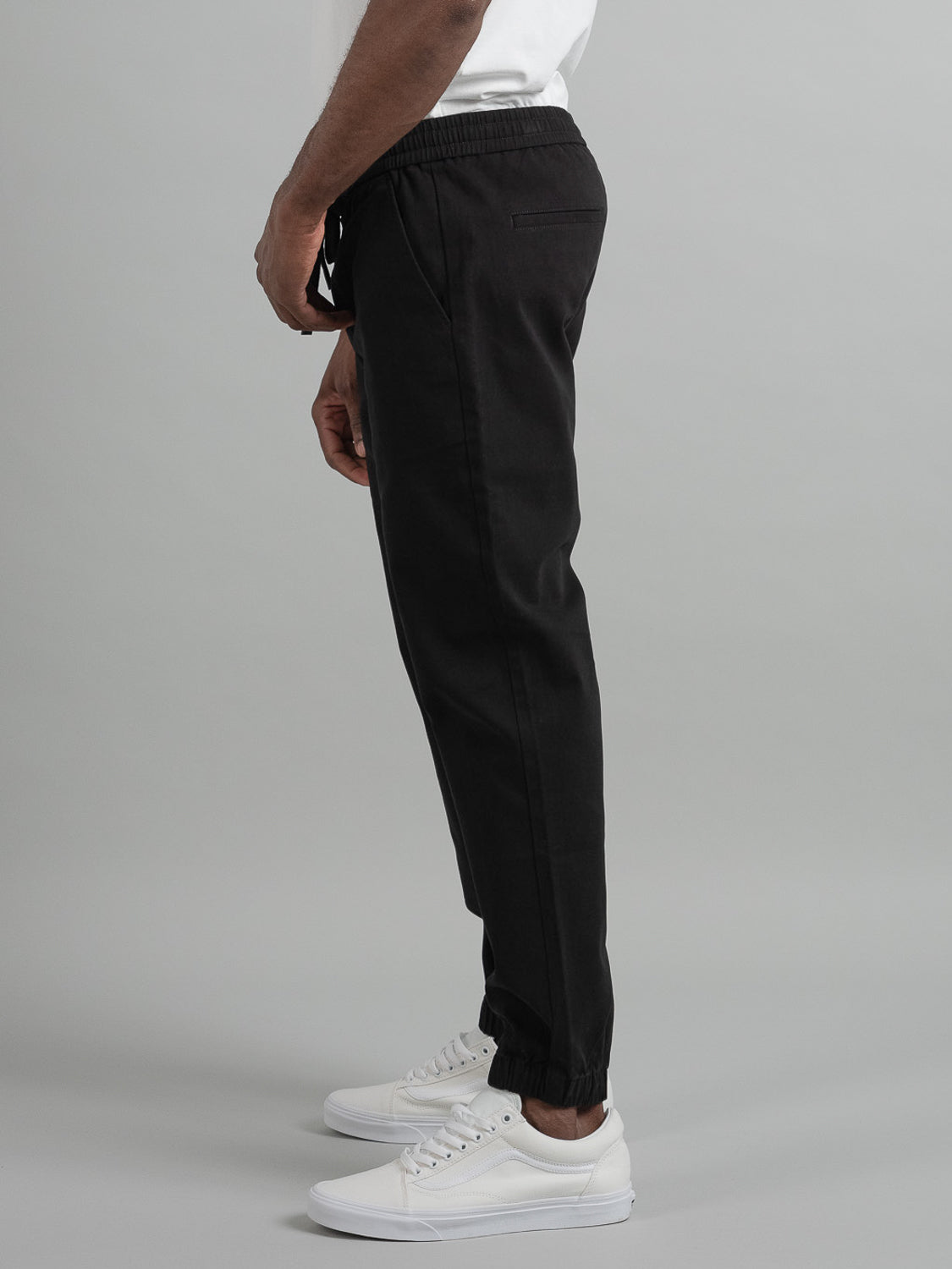 A person in a white T-shirt stands sideways, highlighting ID Menswears Dyker Cotton Twill Drawstring Jogger in black and white sneakers against a plain gray backdrop.