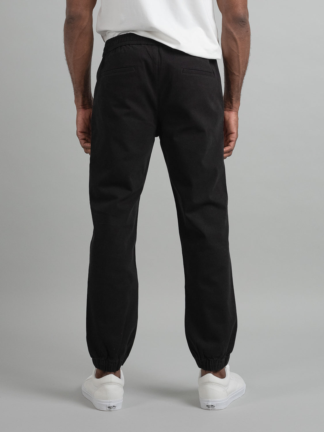 A person is seen from behind wearing a white t-shirt, ID Menswear Dyker Cotton Twill Drawstring Jogger pants in black with elastic waist and cuffs, and white sneakers. The background features a plain light gray color.