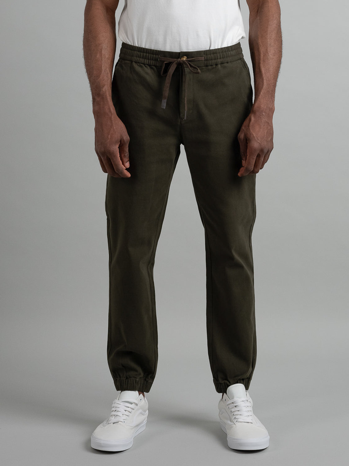A person wears the Dyker Cotton Twill Drawstring Jogger by ID Menswear in olive green, paired with white sneakers. These joggers have an elastic waist for comfort. The background is plain gray, and the persons head is not visible.