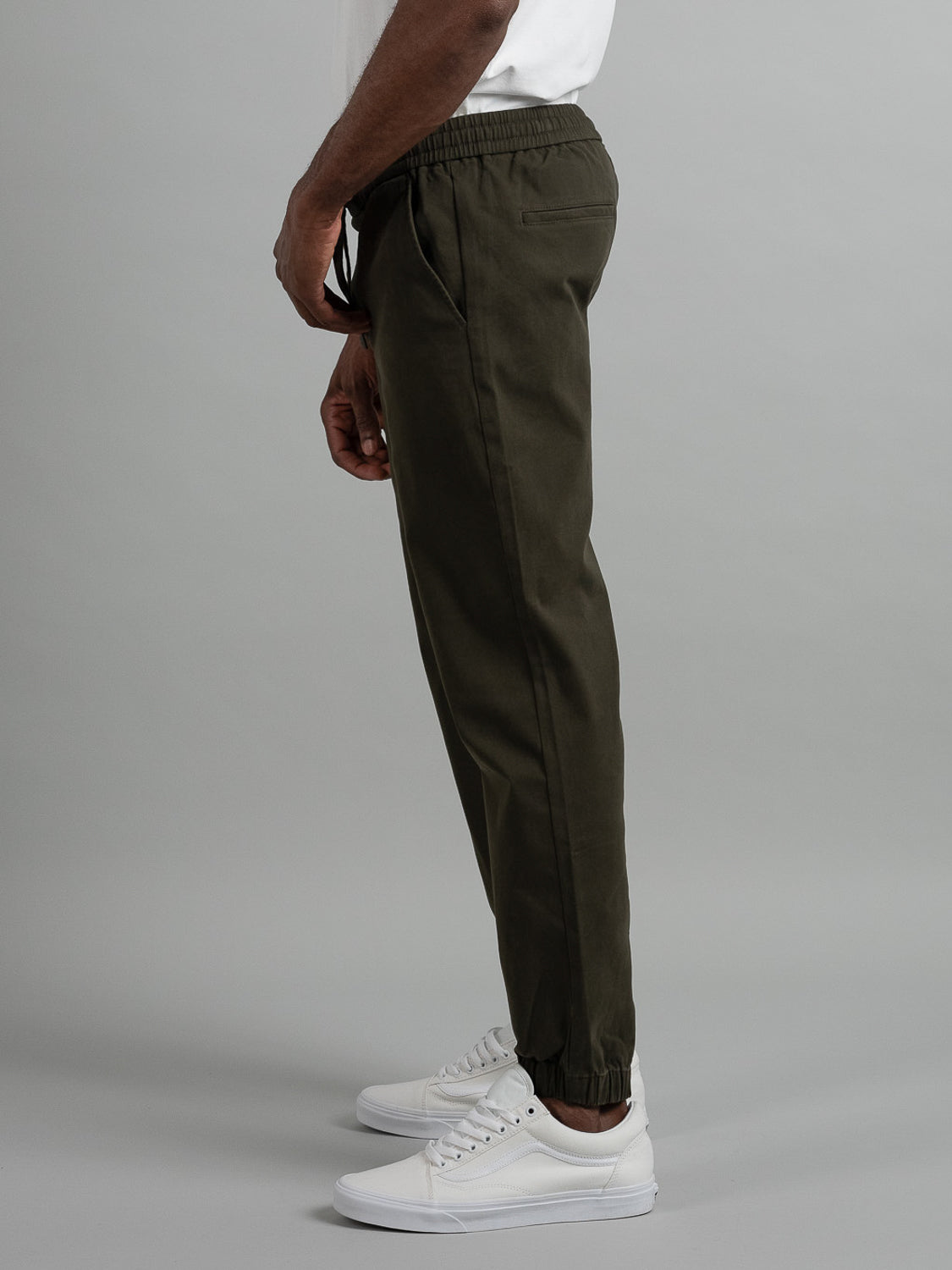 A person in profile against a neutral background wears ID Menswears Dyker Cotton Twill Drawstring Jogger in dark green with an elastic waistband and cuffed ankles, styled with white sneakers. Their hands rest calmly at their sides.