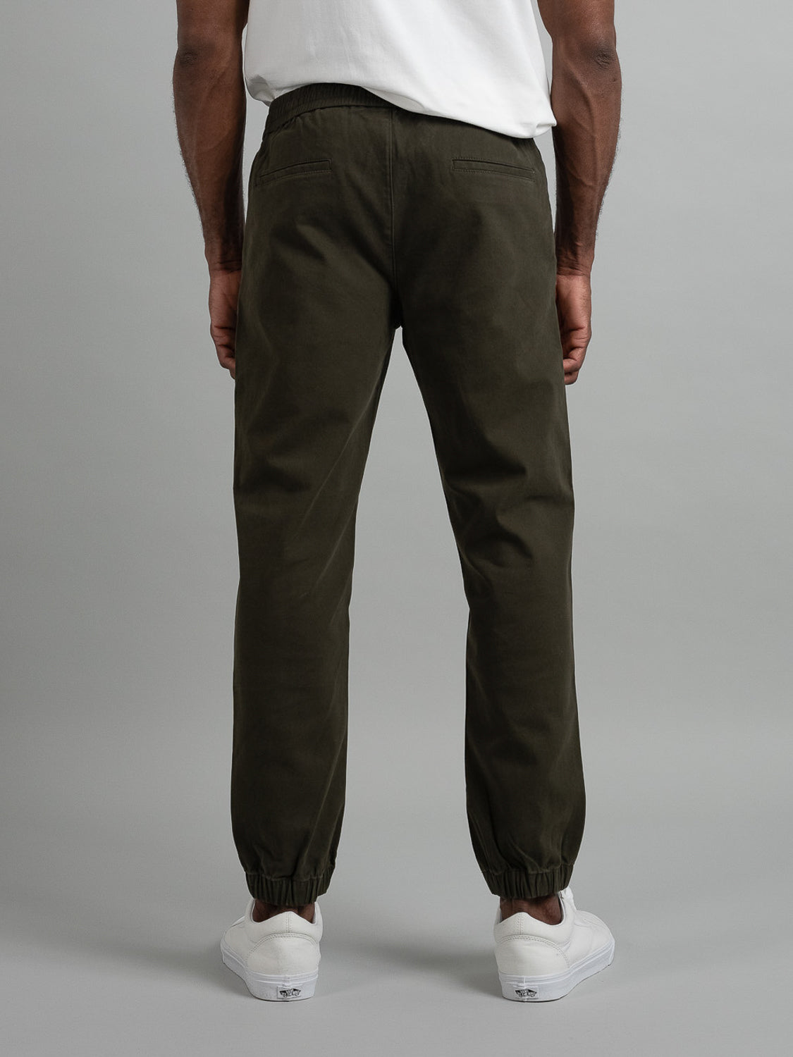 Rear view of a person in a white T-shirt and Dyker Cotton Twill Drawstring Jogger by ID Menswear, in dark olive green with elastic cuffs, standing against a gray background. The joggers fabric enhances the casual vibe, complemented by white sneakers.