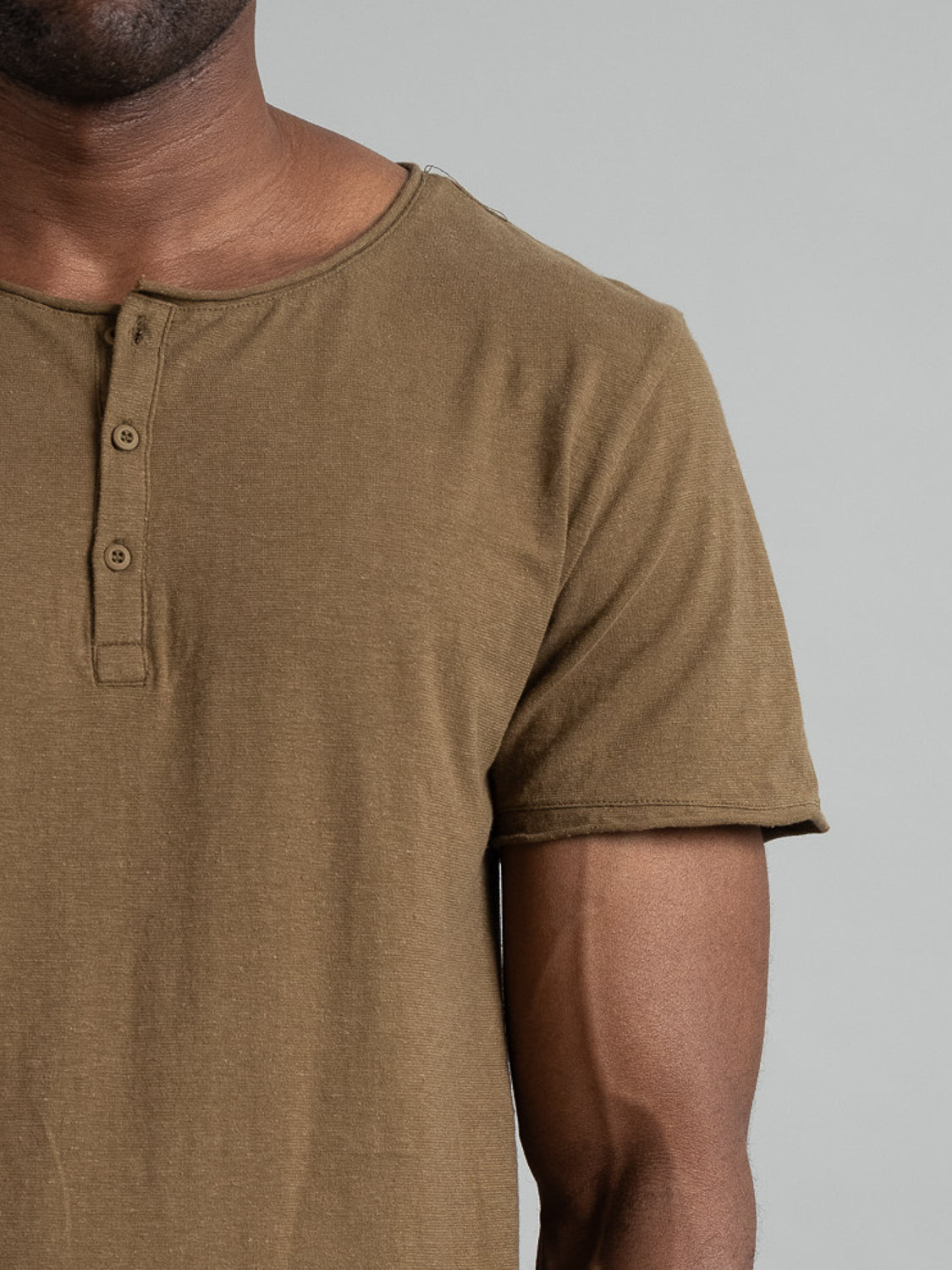 A person models an olive green Legend Hemp Cotton Rolled Neck Henley from ID Menswear, made of organic cotton with a buttoned placket. The breathable henleys comfortable fit is displayed against a plain gray background, showing the upper body and part of the arm.