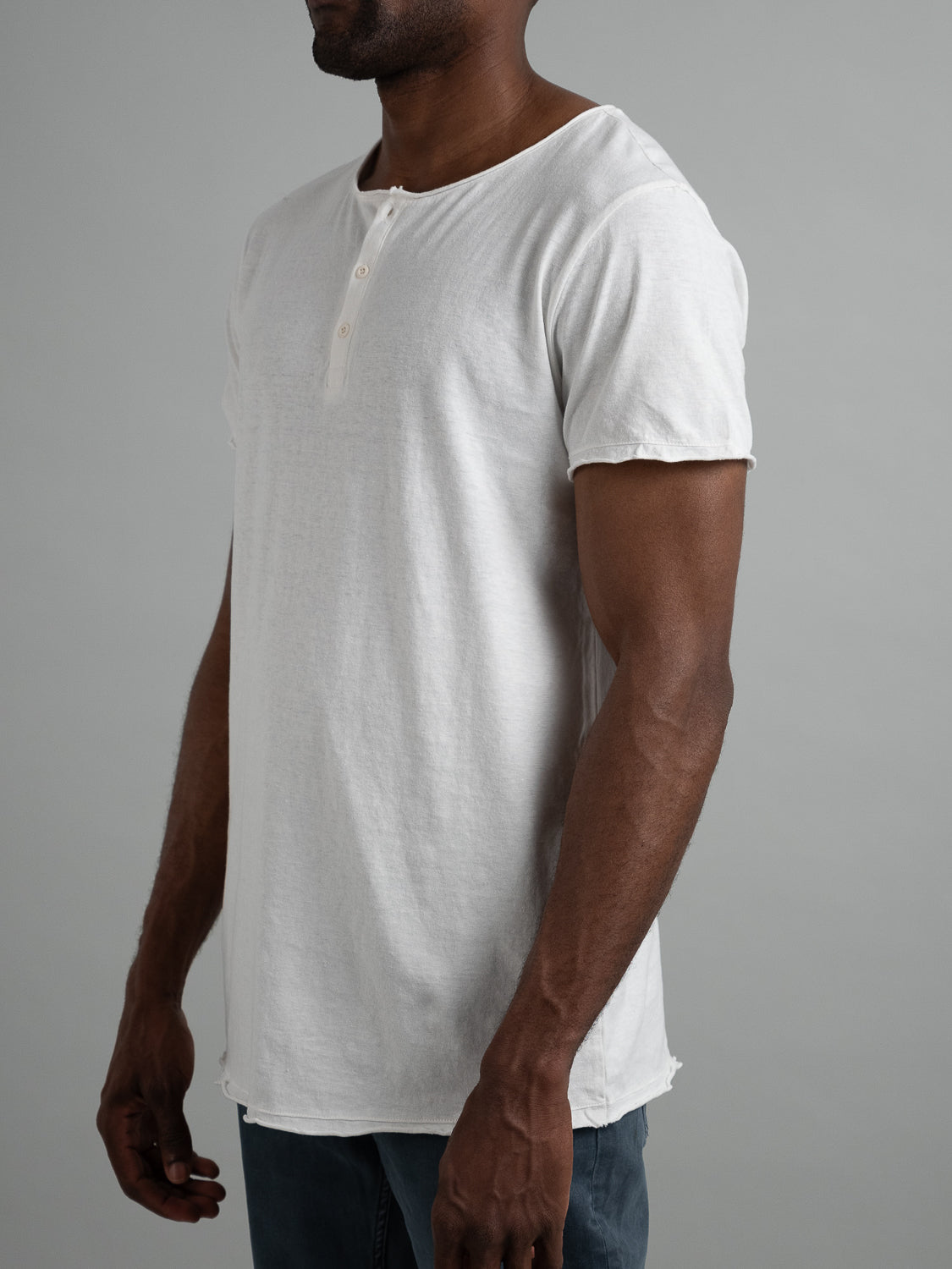 Dressed in ID Menswears Legend Hemp Cotton Rolled Neck Henley, a stylish individual showcases effortless comfort and style. The breathable, organic cotton henley with a three-button placket stands out against dark pants and a plain gray background.