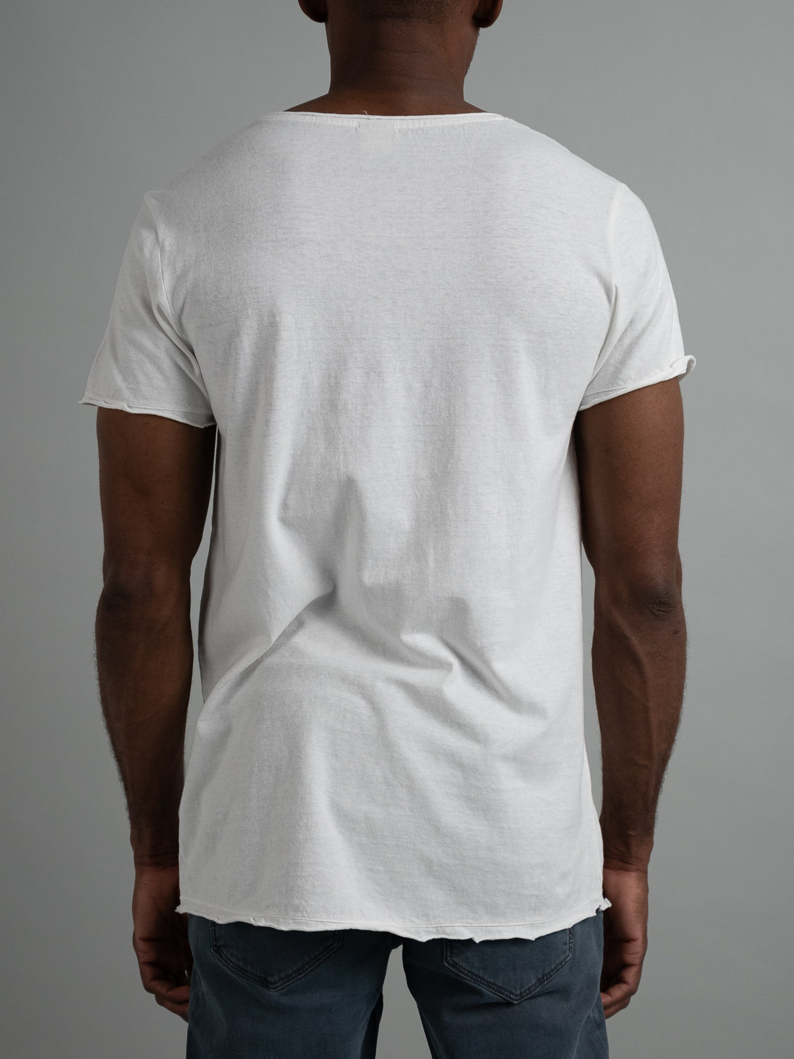 A person stands with their back to the camera, wearing an ID Menswear Legend Hemp Cotton Rolled Neck Henley and dark jeans. The neutral gray backdrop emphasizes their naturally simple style.