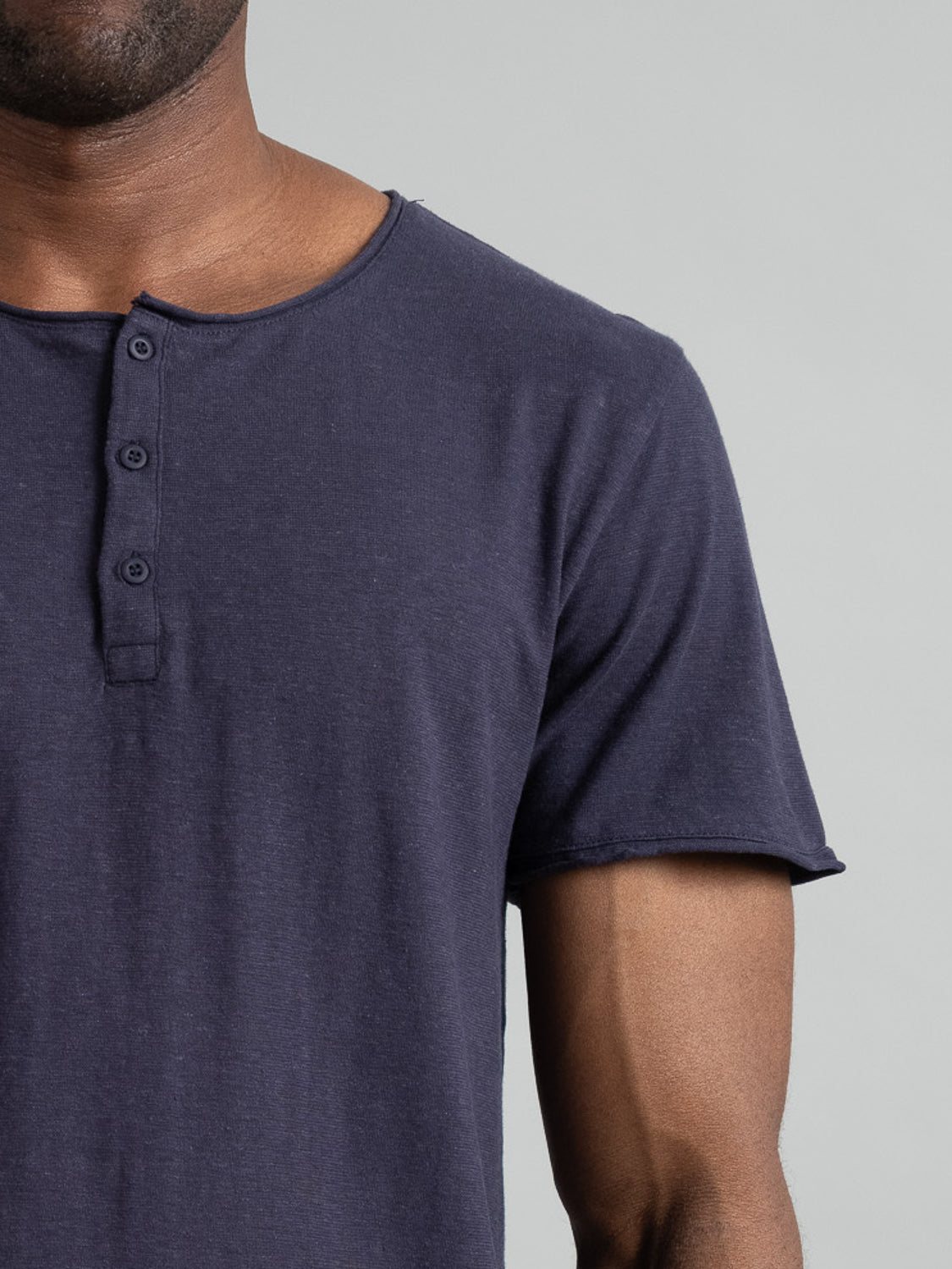 A person is shown in a cropped photo wearing the Legend Hemp Cotton Rolled Neck Henley by ID Menswear. The shirt is dark blue, short-sleeved, and made from breathable organic cotton with three buttons. The background is plain and only the upper torso is visible.