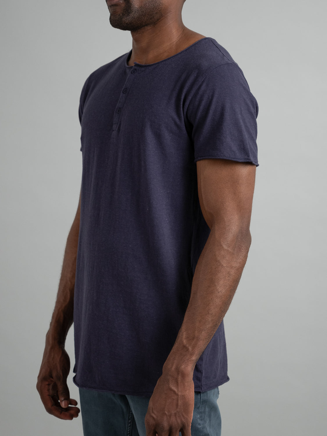 A person models the dark blue Legend Hemp Cotton Rolled Neck Henley by ID Menswear against a grey backdrop. The textured fabric and buttoned neckline of this organic cotton henley highlight its natural comfort.
