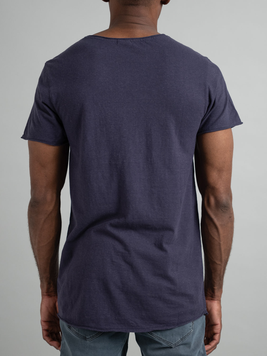 A person wearing a dark blue Legend Hemp Cotton Rolled Neck Henley from ID Menswear and light blue jeans stands with their back to the camera. They have short hair and are set against a neutral gray backdrop.