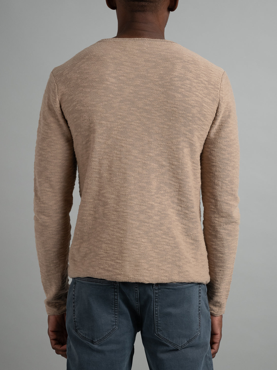 A person in a beige ID Menswear Havana Slub Yarn Sweater Tee and blue jeans faces away from the camera, set against a plain gray backdrop. The sweaters textured pattern offers a premium feel, ideal for casual wear.