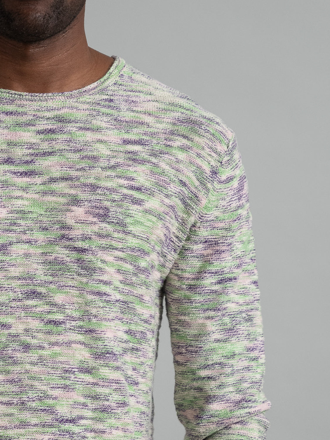 Close-up of an individual displaying a Havana Slub Yarn Sweater Tee from ID Menswear, featuring green, purple, and white tones. This premium casual garment pops against the simple gray background, revealing just the upper torso and a portion of an arm.