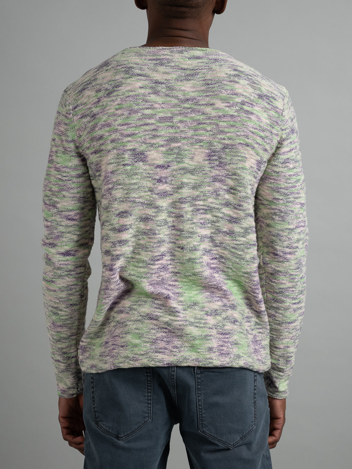 A person in an ID Menswear Havana Slub Yarn Sweater Tee, showcasing green, purple, and white tones, pairs it with blue jeans for a premium yet casual look while posing against a gray background.