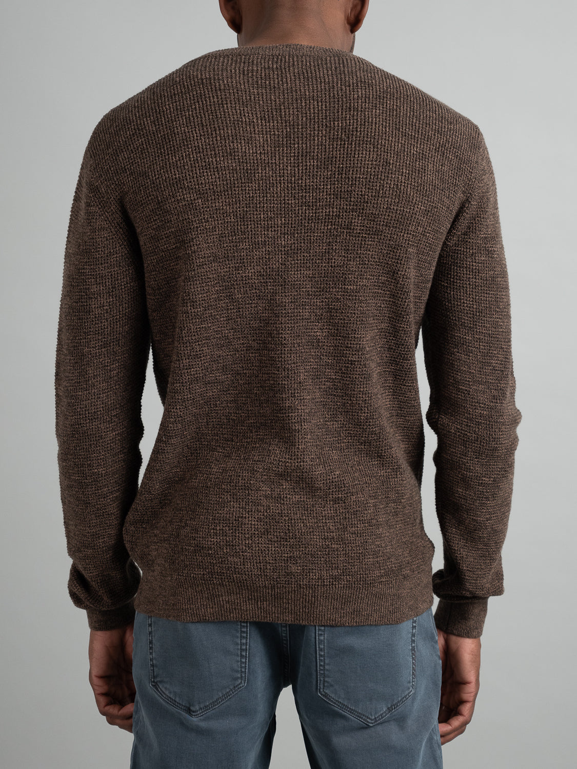 Wearing the Oslo Waffle Knit Sweater by ID Menswear and light blue jeans, a person is facing away from the camera against a plain gray background, embodying relaxed casual wear.