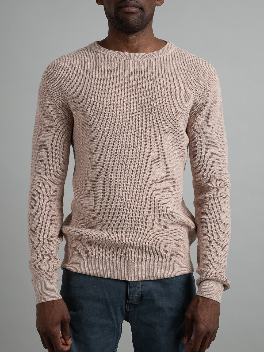 Against a plain gray backdrop, a person in an Oslo Waffle Knit Sweater by ID Menswear and dark pants stands casually with arms at their sides. The sweaters textured cotton pattern adds depth, epitomizing effortless casual wear.