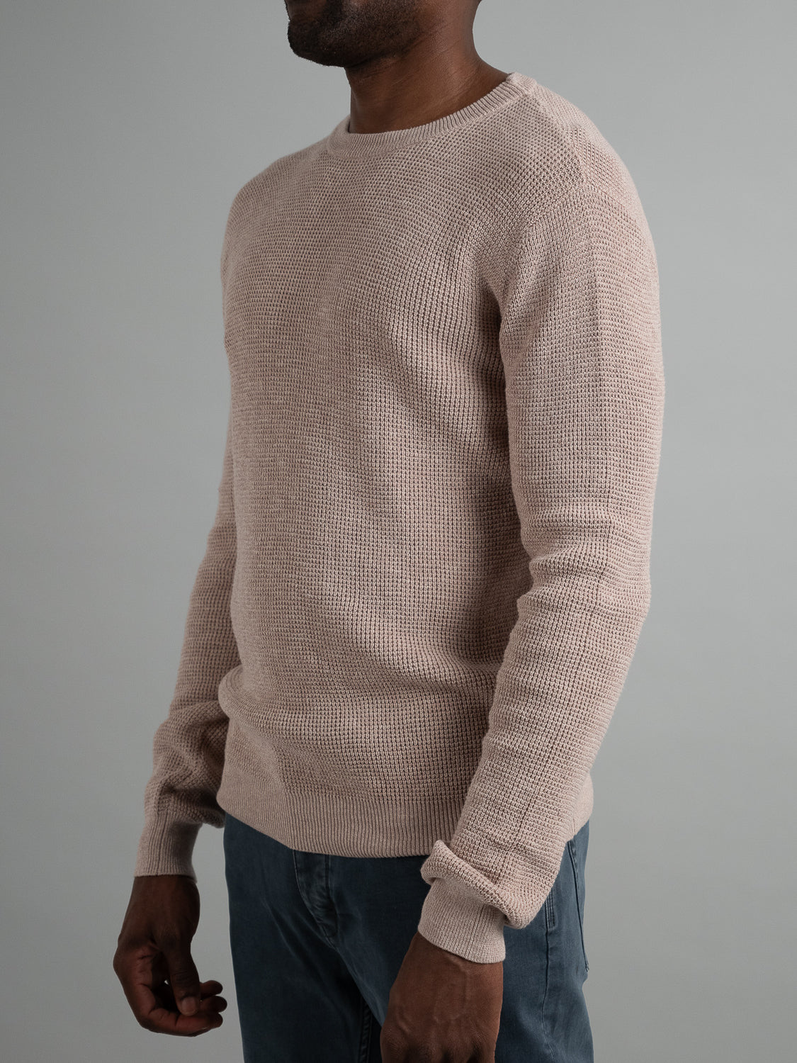 Wearing the beige Oslo Waffle Knit Sweater by ID Menswear against a gray backdrop, a person showcases the sweaters texture and fit. Their face is hidden, and they pair it with dark pants, capturing casual style effortlessly.