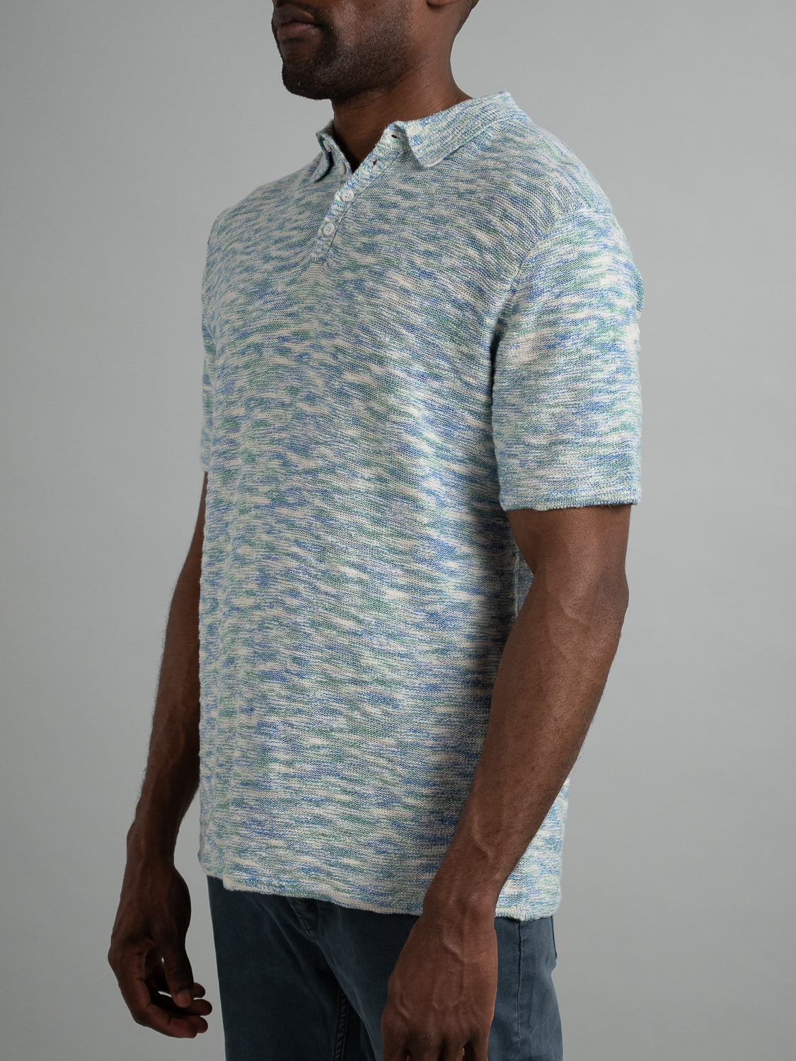 A person wearing ID Menswears Miami Reverse Knit Cotton Polo in light blue and green, featuring a collar and button placket, stands against a plain background. The breathable, marled cotton-knit shirt is paired with dark pants for a casual look.