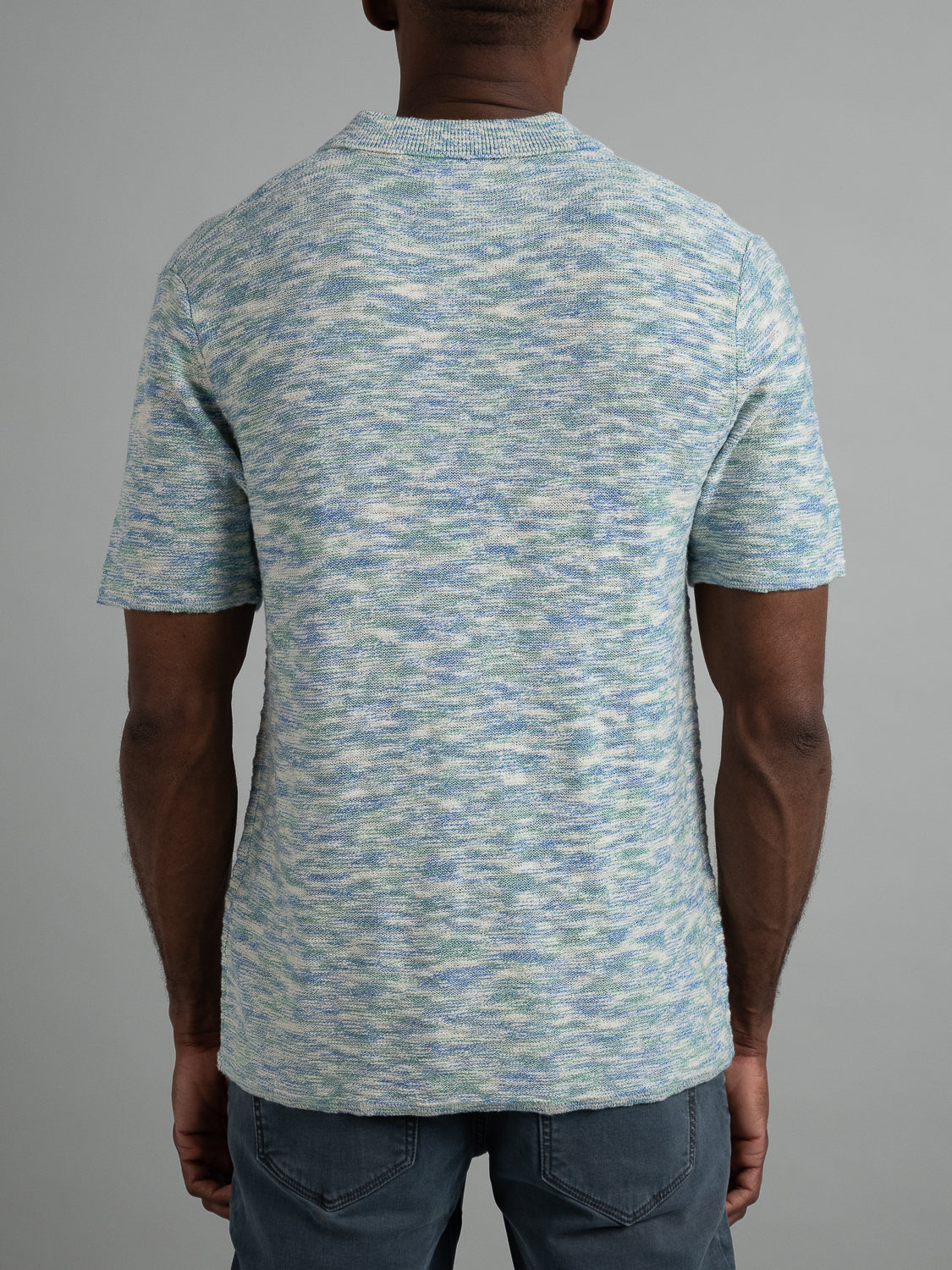 Rear view of a person wearing the Miami Reverse Knit Cotton Polo by ID Menswear, featuring a light blue and white marbled design for breathable comfort, with their arms relaxed at their sides against a gray background, paired with casual gray pants.
