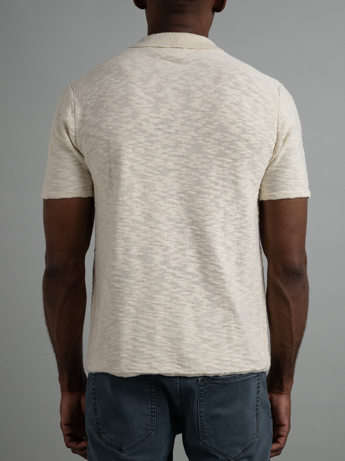 Rear view of a person in an ID Menswear Miami Reverse Knit Cotton Polo and dark jeans, highlighting casual wear with a breathable fit, against a plain gray backdrop.