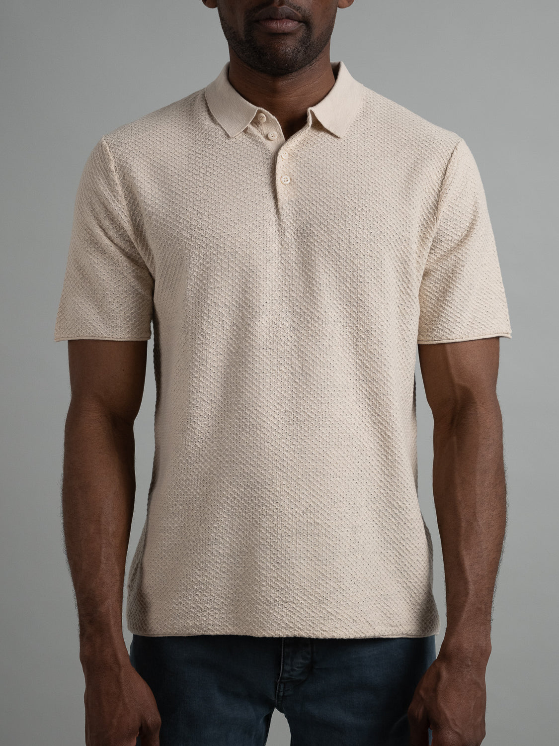 A man models the Phoenix Super Fine Cotton Knit Polo by ID Menswear. The textured beige short-sleeve shirt, featuring a collar and buttons, is paired with dark jeans against a plain gray backdrop, delivering an effortlessly stylish and versatile casual look with his arms at his sides.