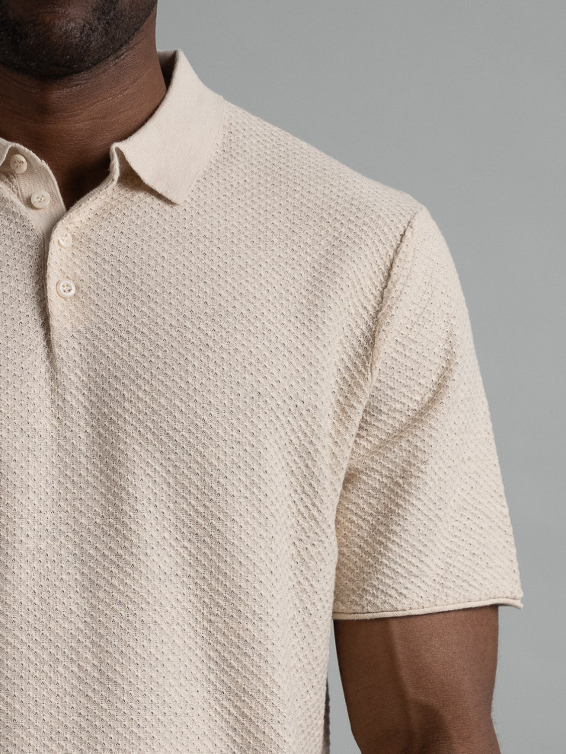 A person wearing the Phoenix Super Fine Cotton Knit Polo from ID Menswear stands against a gray background. This textured beige polo, ideal for casual wear, has short sleeves and three collar buttons, featuring a small raised diamond pattern for a versatile stylish look.