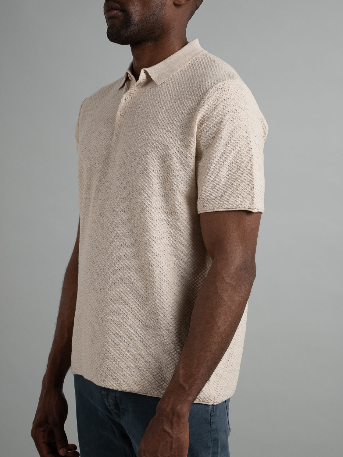 An individual models the Phoenix Super Fine Cotton Knit Polo by ID Menswear, set against a plain gray backdrop. The shirts textured beige design elevates its casual allure. Relaxed arms and dark pants complete their versatile, stylish look.