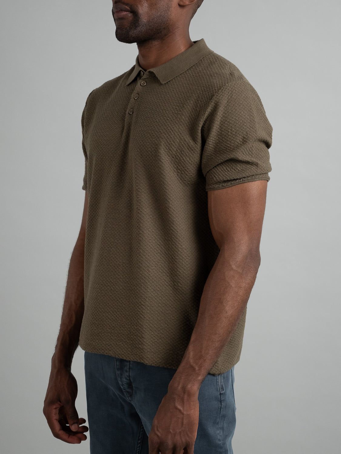 A man in an ID Menswear Phoenix Super Fine Cotton Knit Polo, featuring textured olive green cotton-knit, and blue jeans stands against a grey background. His arms are at his sides, presenting a versatile look ideal for casual wear, with his face unseen.
