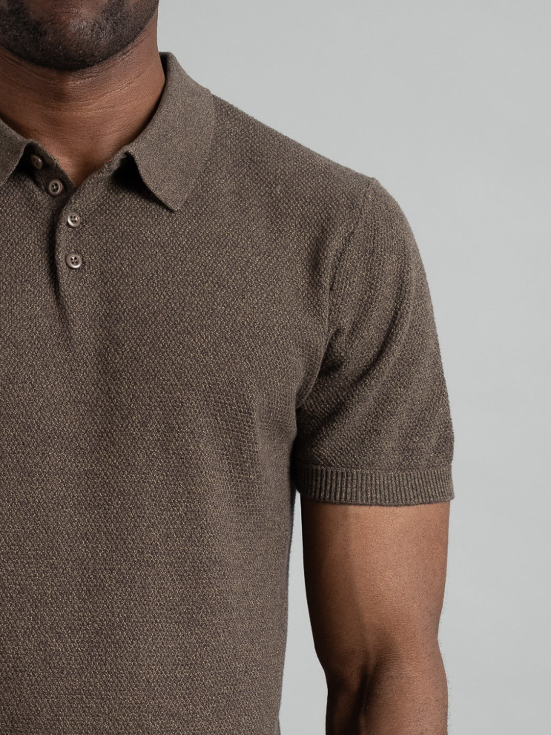 An individual dons an ID Menswear Austin Knitted Cotton Polo, featuring a brown textured knit with a collar and buttons. Photographed from neck to waist against a plain gray background, the image highlights the shirts casual style along with part of one arm.