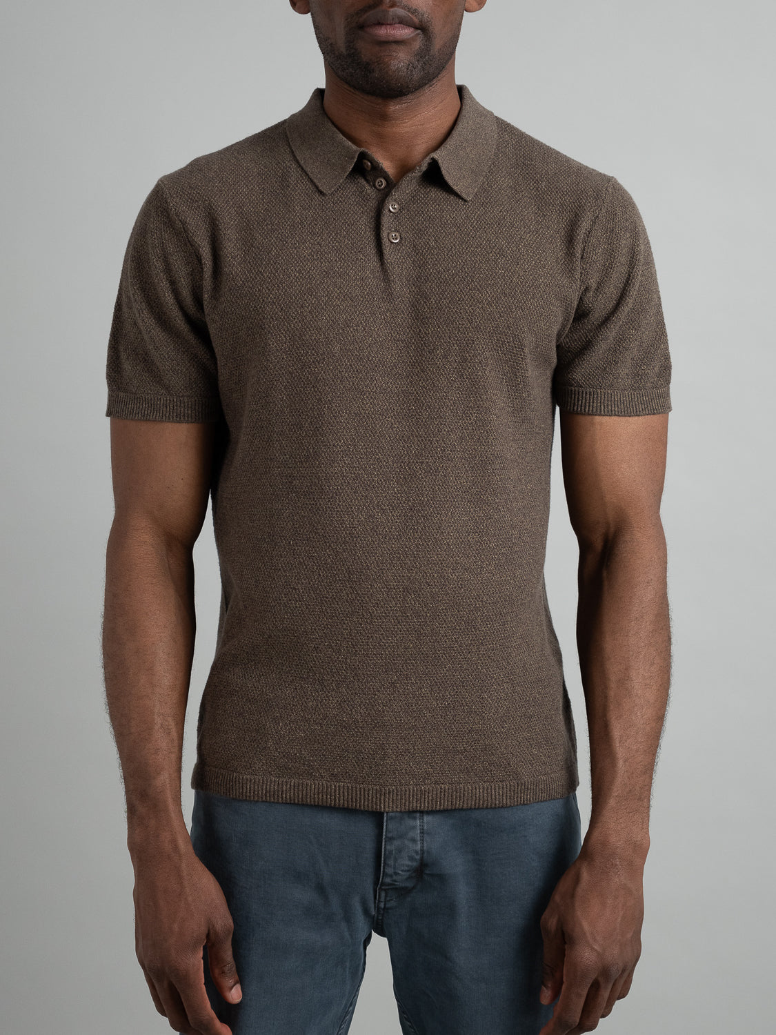 A person wears the Austin Knitted Cotton Polo by ID Menswear, featuring a textured brown cotton-knit and short sleeves, paired with blue jeans. They stand with arms relaxed against a plain light gray background, enjoying the breathable comfort.