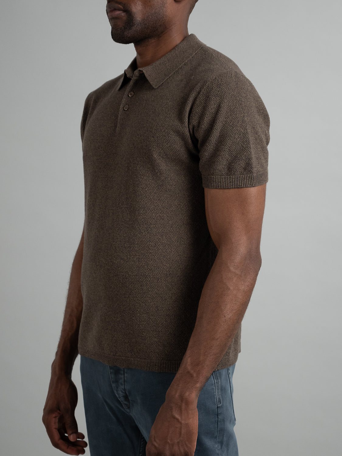 A person models the ID Menswear Austin Knitted Cotton Polo in brown against a plain gray backdrop, paired with blue jeans. Emphasizing the breathable fit on the upper body, their face is obscured for a focus on casual elegance.