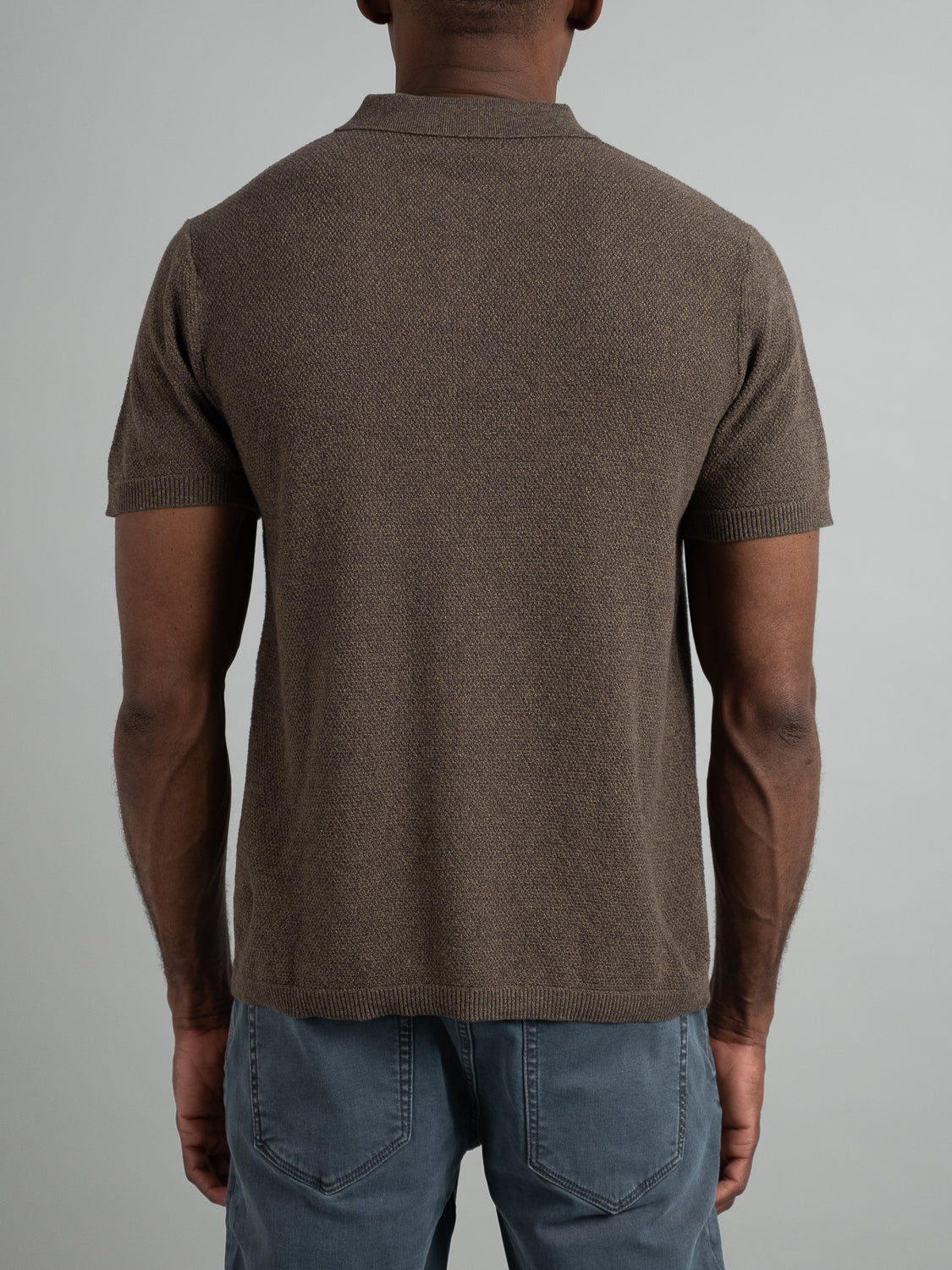 A person wearing ID Menswears Austin Knitted Cotton Polo in breathable brown fabric paired with blue jeans, facing away from the camera against a neutral background.