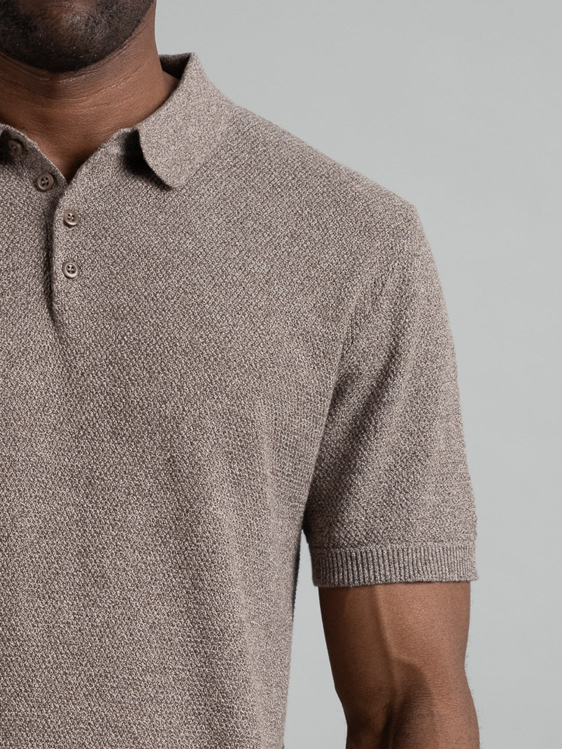A person is seen from the chest to shoulder in an Austin Knitted Cotton Polo by ID Menswear, featuring a textured brown cotton-knit that offers a breathable fit. Shot against a gray backdrop, the image subtly highlights the understated elegance of this casual piece. Face and arm partially visible.