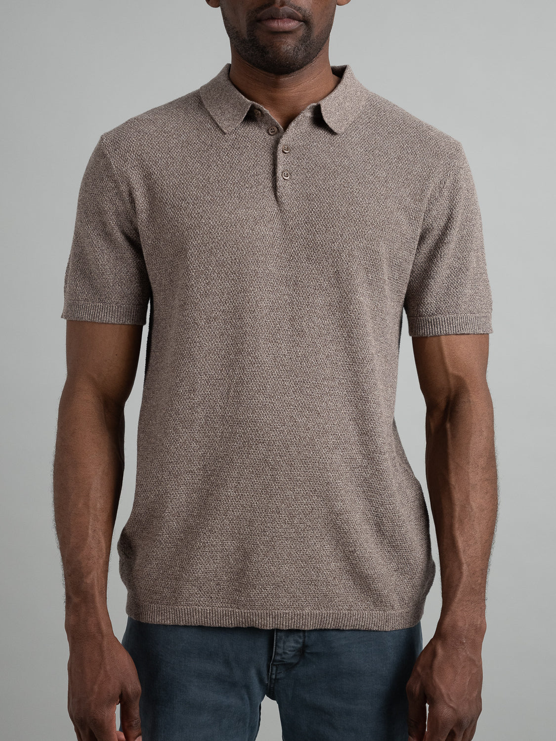 A man in the Austin Knitted Cotton Polo by ID Menswear, featuring a textured beige design with a classic collar and button placket, stands against a gray backdrop. He pairs it perfectly with dark gray pants, creating an ideal mix of casual style and breathability.