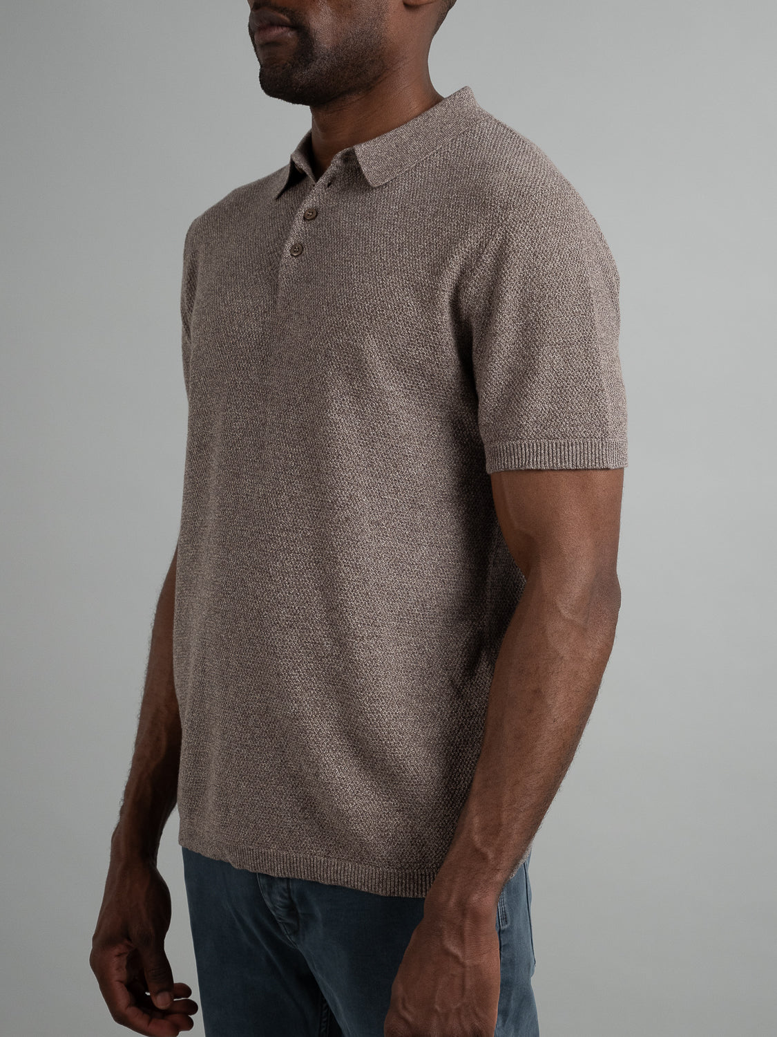 A person models ID Menswears Austin Knitted Cotton Polo, a short-sleeve, light brown shirt with a breathable fit and three-button placket, perfectly paired with dark jeans against a plain gray backdrop for a casual look.