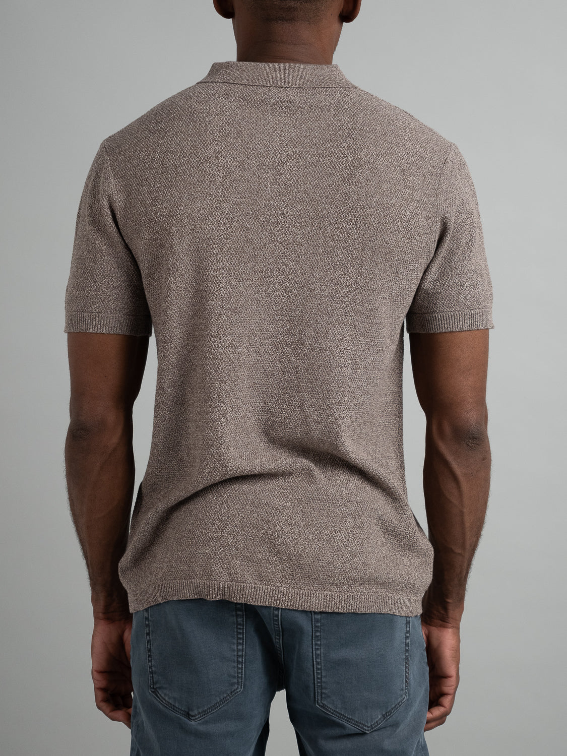 A person is pictured from the back wearing the Austin Knitted Cotton Polo by ID Menswear in beige, paired with blue jeans against a gray background, showcasing a breathable fit.