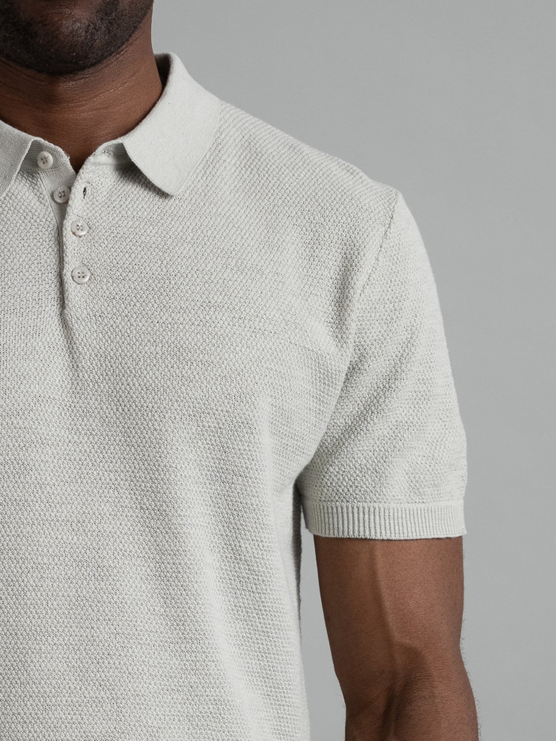 A person in a light gray Austin Knitted Cotton Polo from ID Menswear is shown from the shoulders down. The shirt features short sleeves and a buttoned collar, ideal for casual wear with its breathable fit against a muted gray background.