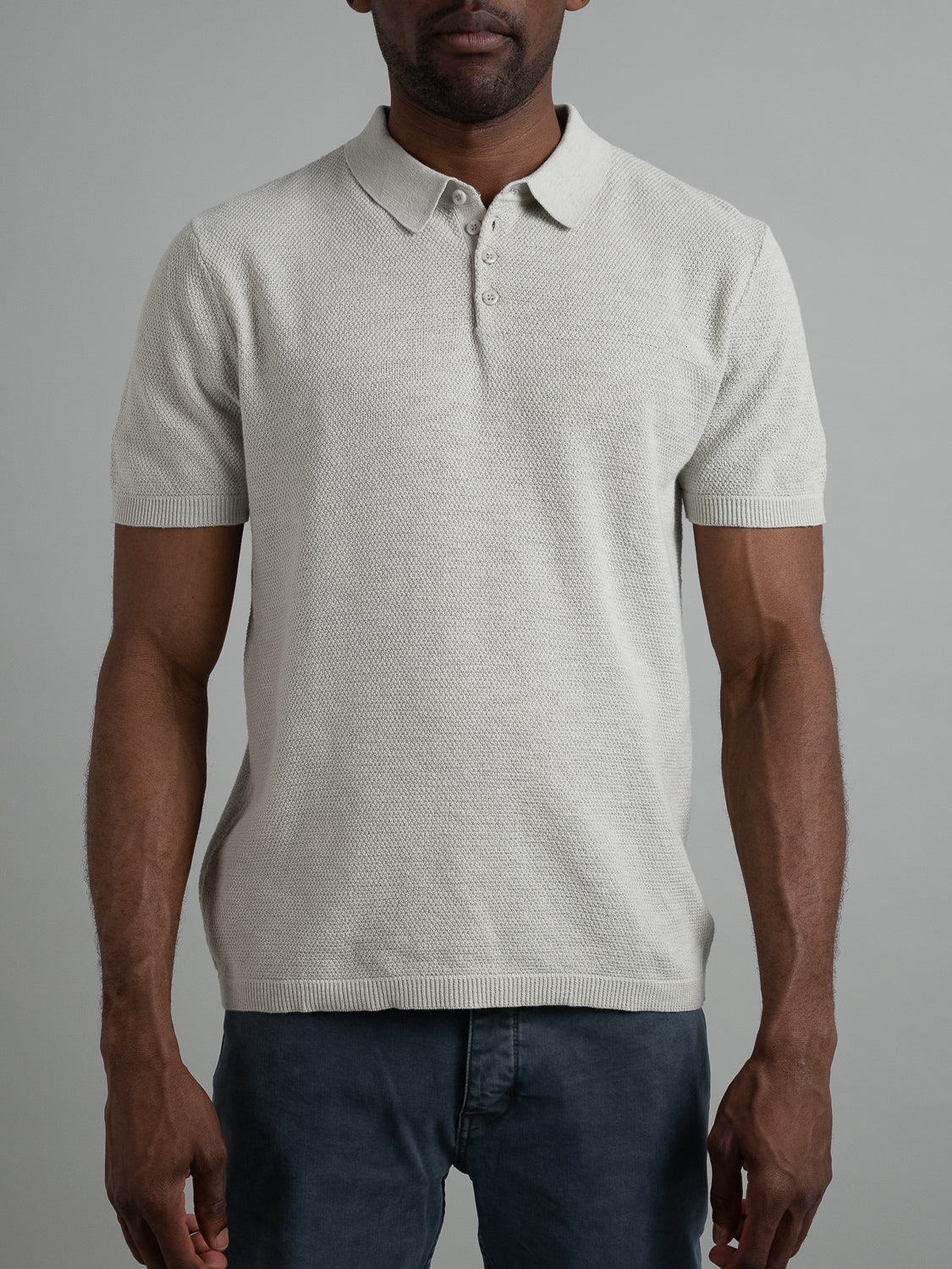 Someone in dark pants stands against a neutral background wearing the Austin Knitted Cotton Polo by ID Menswear. This light gray, short-sleeve polo features a textured fabric and buttoned collar, offering a comfortable and breathable fit perfect for casual wear.