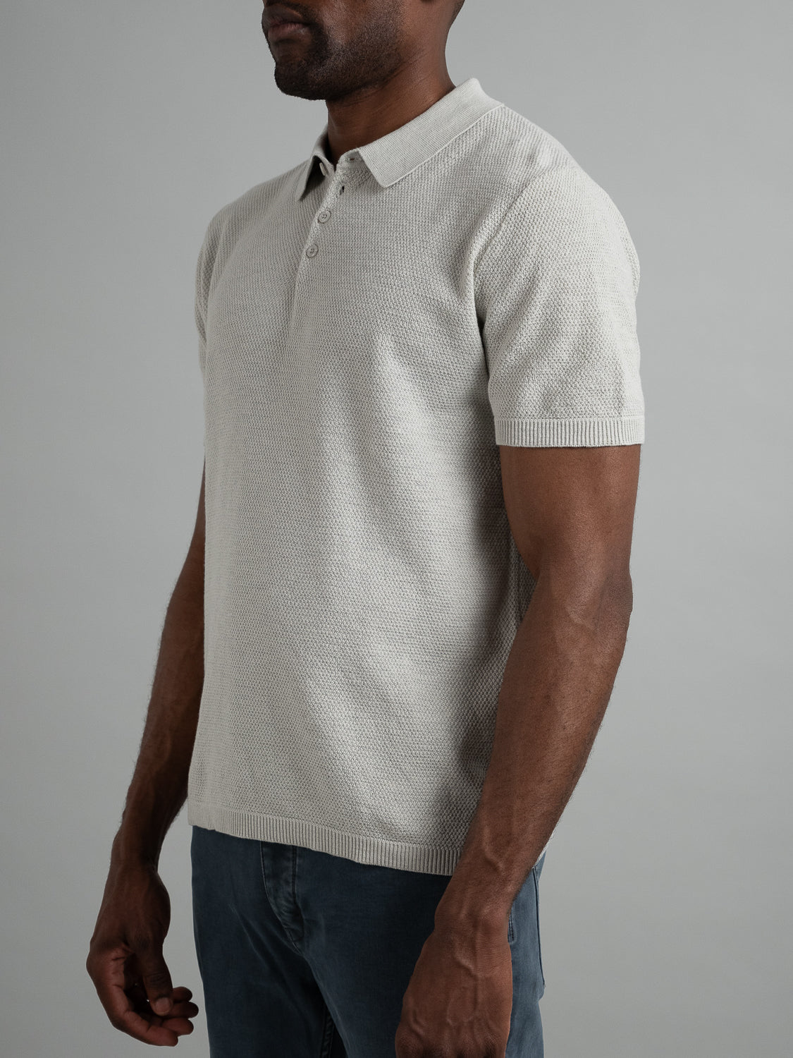 A person wearing an Austin Knitted Cotton Polo by ID Menswear stands against a plain gray background. Their arms are relaxed by their sides, paired with dark pants. The focus is on the breathable fit and texture of this light gray short-sleeve shirt, embodying effortless casual wear.
