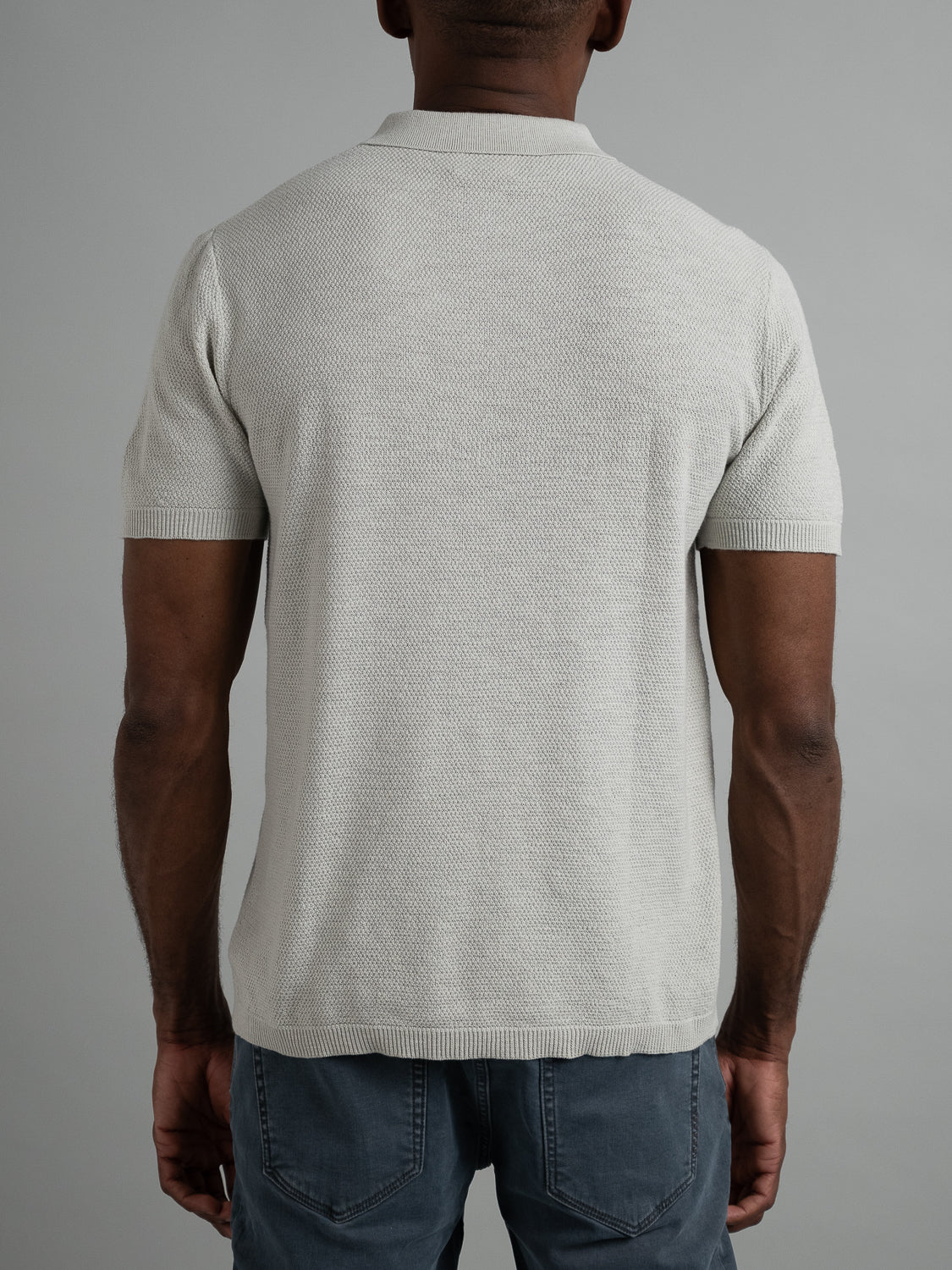 Rear view of a person wearing ID Menswears Austin Knitted Cotton Polo in light gray, paired with blue jeans. Standing against a gray backdrop, their muscular build highlights the polo’s breathable fit as their arms rest relaxed by their sides.